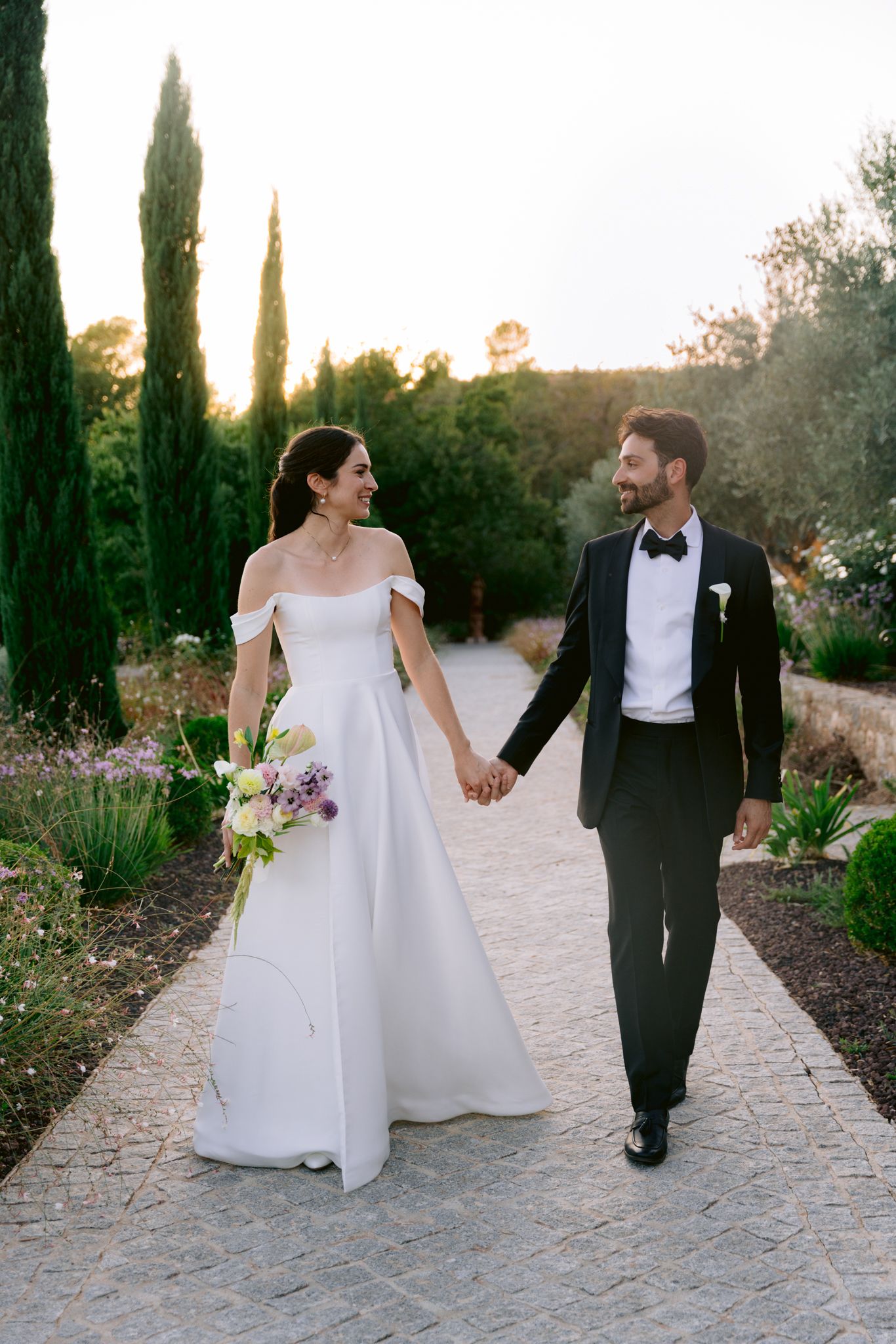 Newlyweds walk hand in hand along a stone path lined with cypress trees during a romantic sunset photoshoot.