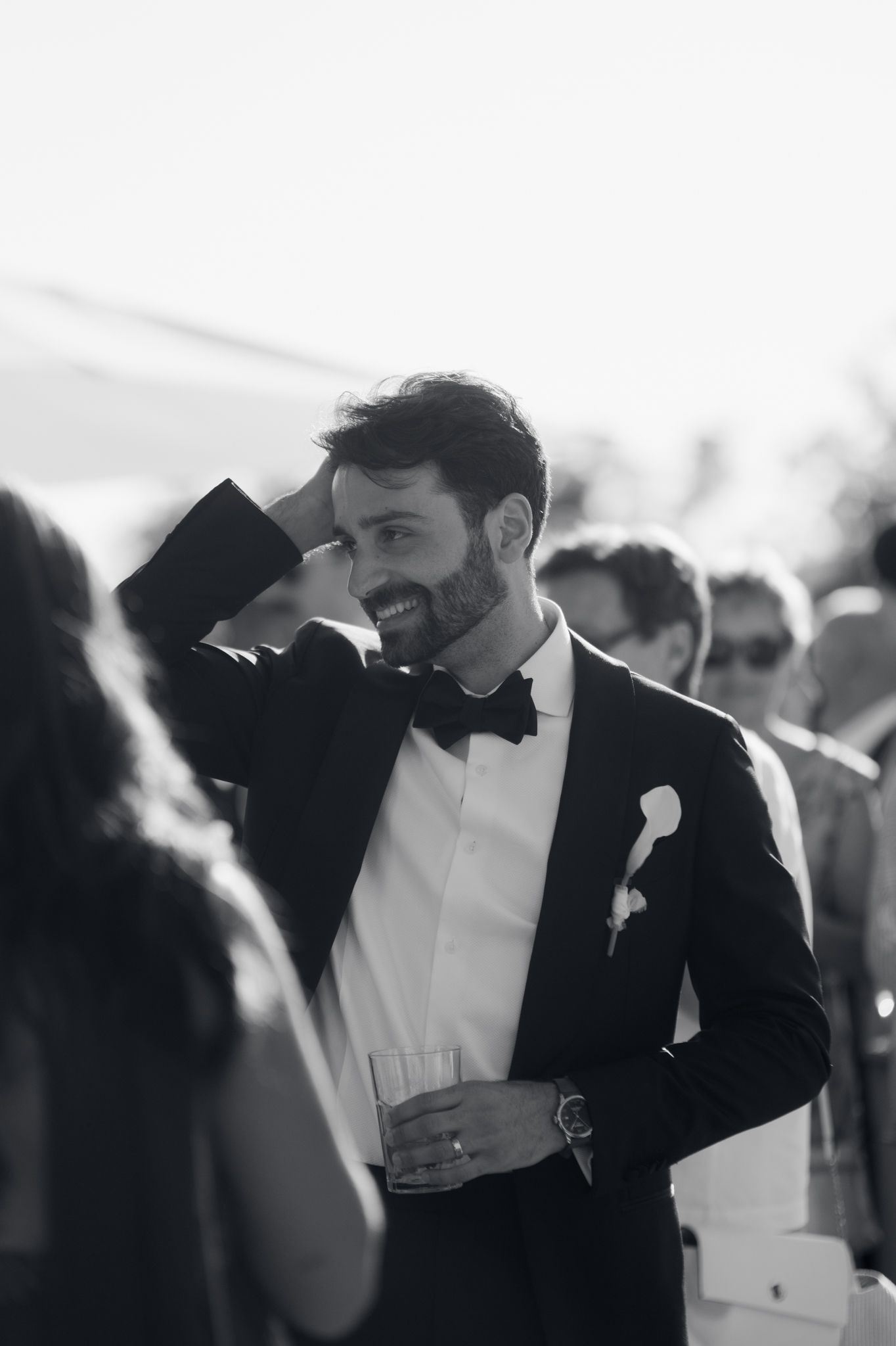 Black and white candid wedding photo showing groom in tuxedo with boutonniere laughing during reception.