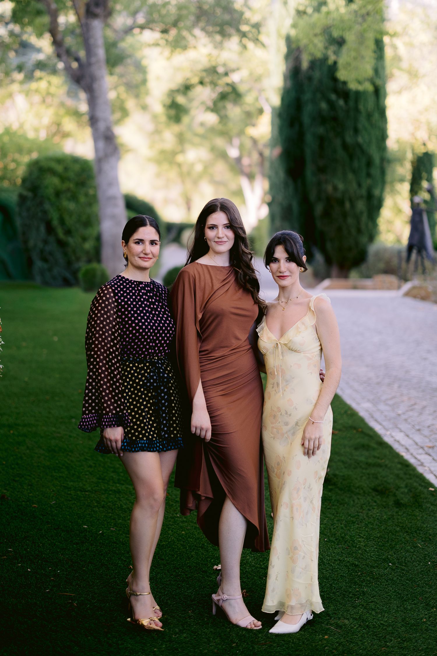 Three women in elegant dresses pose together on green lawn at outdoor garden event.
