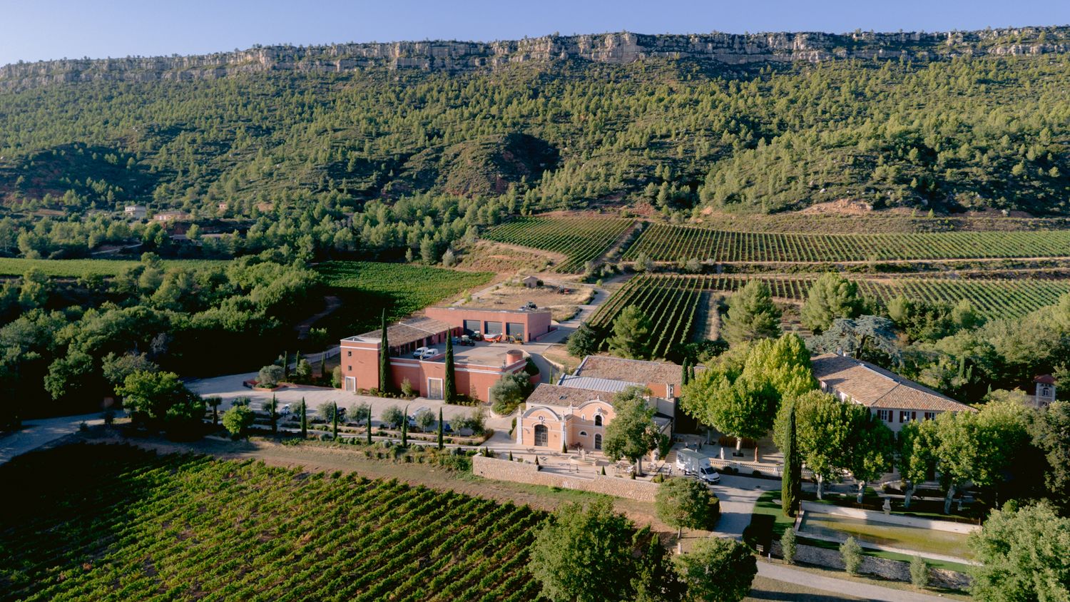 Scenic aerial panorama of a Tuscan countryside with rolling hills, vineyards, and historic stone buildings under blue skies.