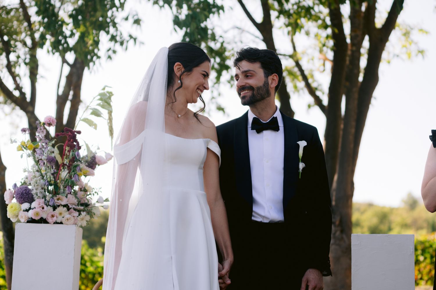 A bride and groom share intimate moments during their outdoor wedding ceremony under lush green trees.