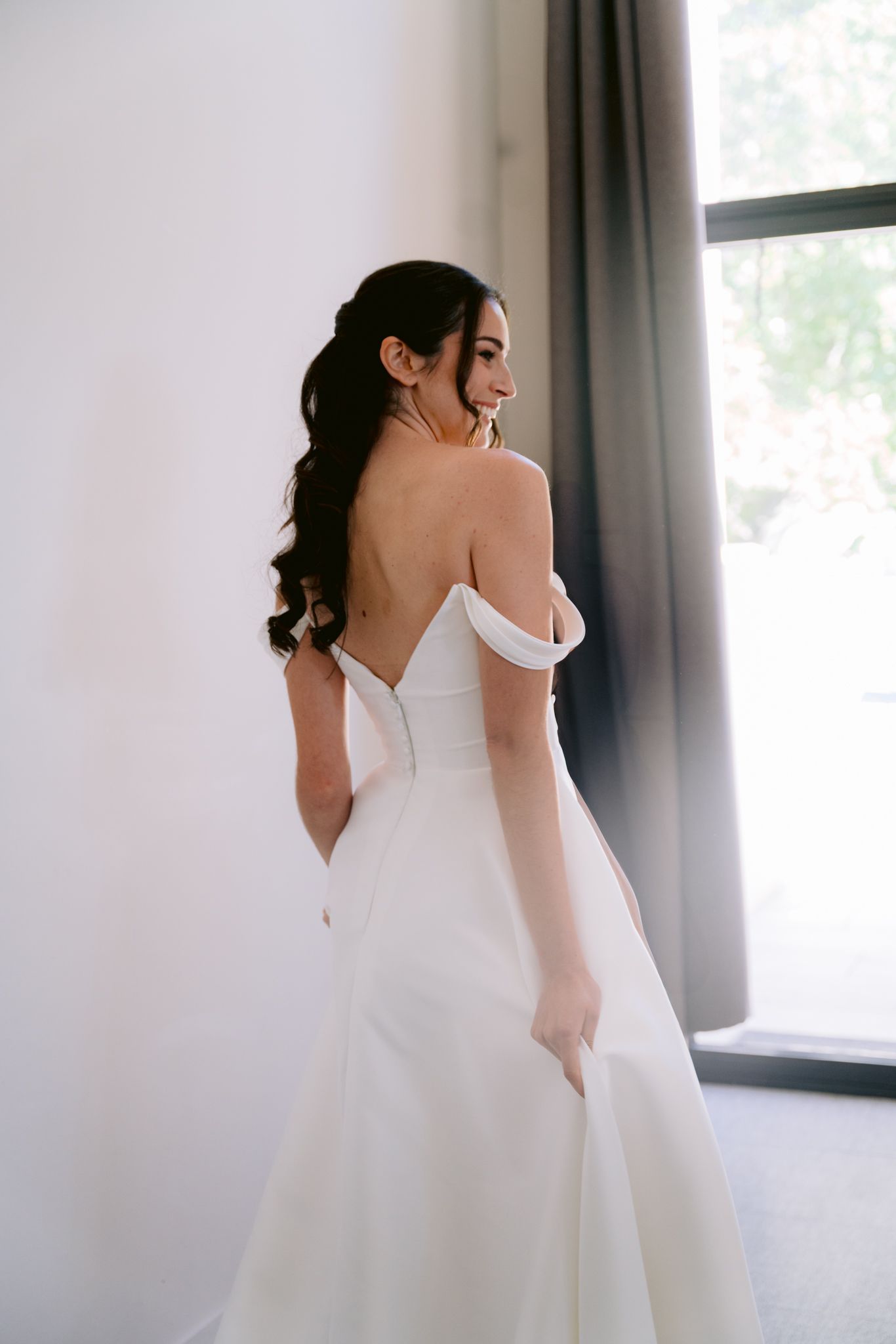 A bride in a white wedding dress with a plunging back design stands elegantly in a softly lit room.