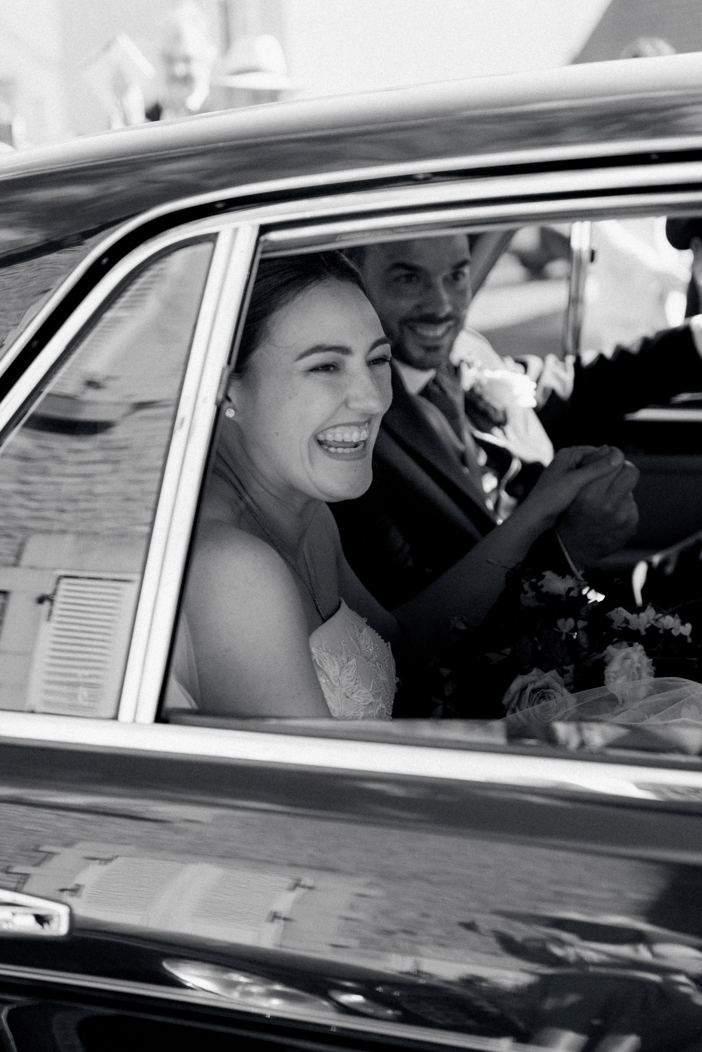 Black and white photo of a laughing bride and smiling groom sitting in a classic wedding car.