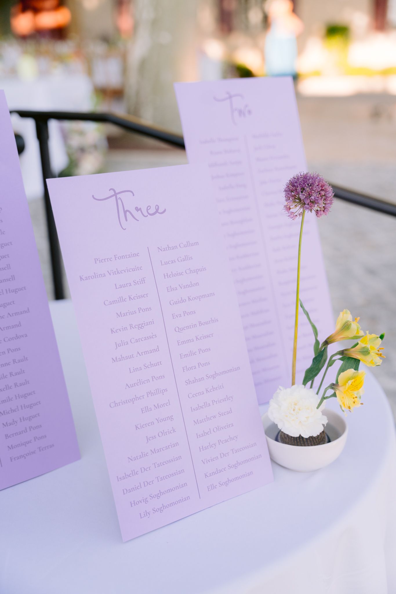 Wedding seating chart cards in lavender purple with floral accents and yellow daffodils on white tablecloth.