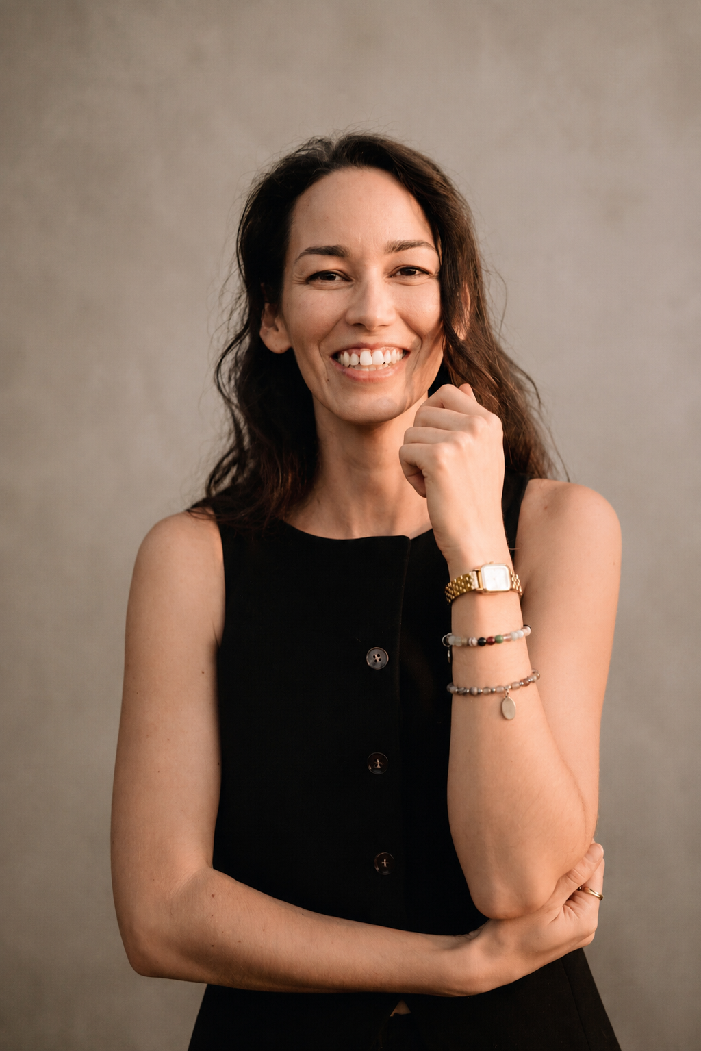 Smiling woman in black sleeveless dress wearing gold watch and bracelets, posing confidently against neutral background.