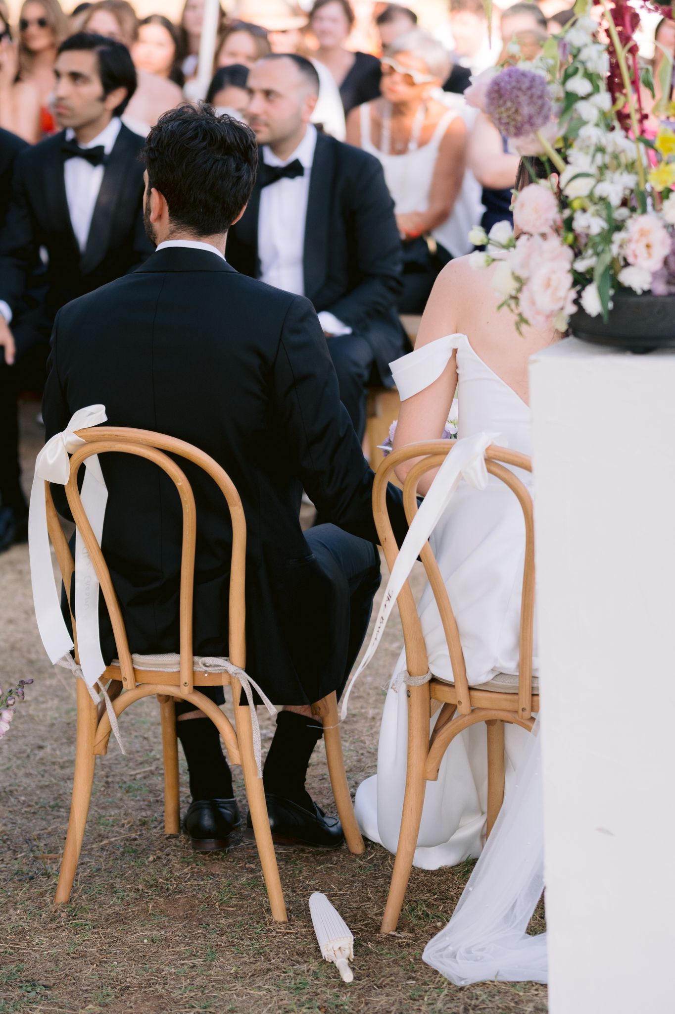 Elegant wedding reception detail showing couple seated in wooden bentwood chairs with white ribbons tied to the backs.