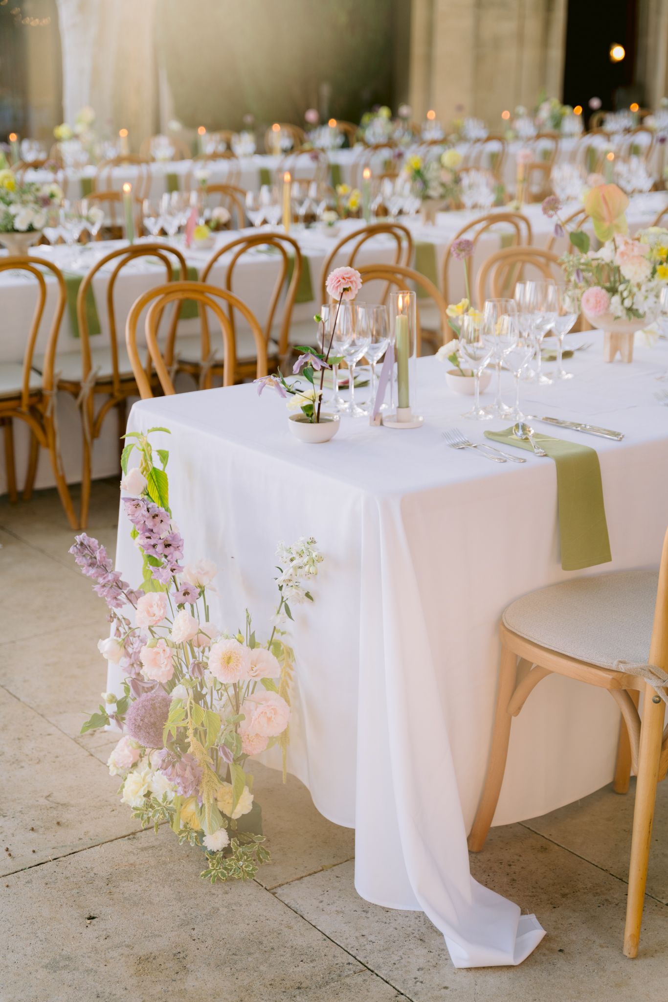 Elegant wedding reception table with white linen, pastel floral arrangements, and wooden bentwood chairs in a softly lit venue.