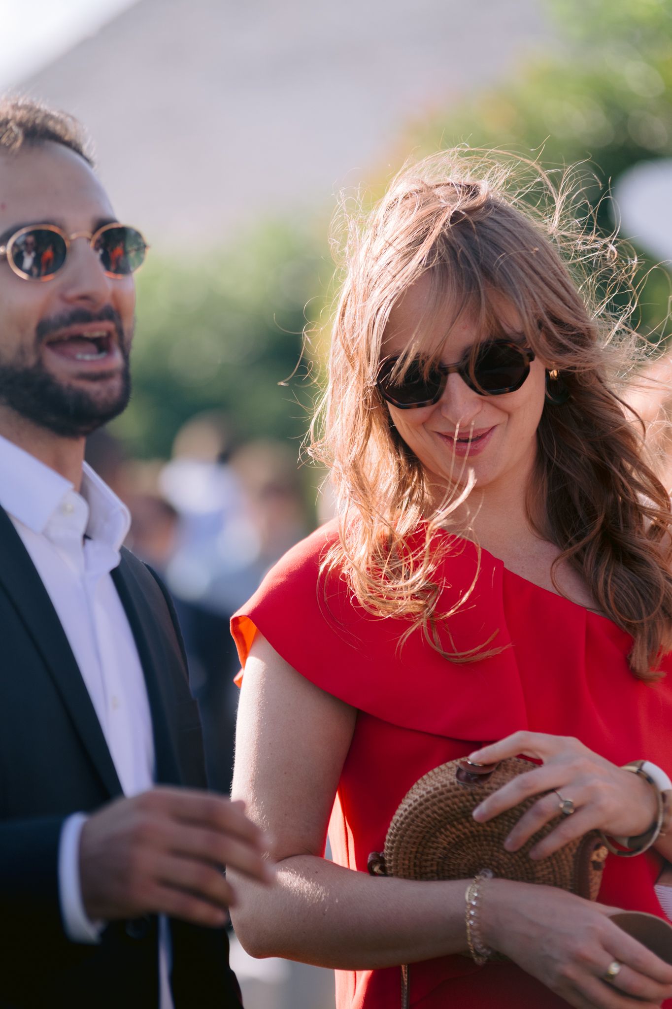 A man in a suit and a woman in a red dress wearing sunglasses socializing outdoors at an event.