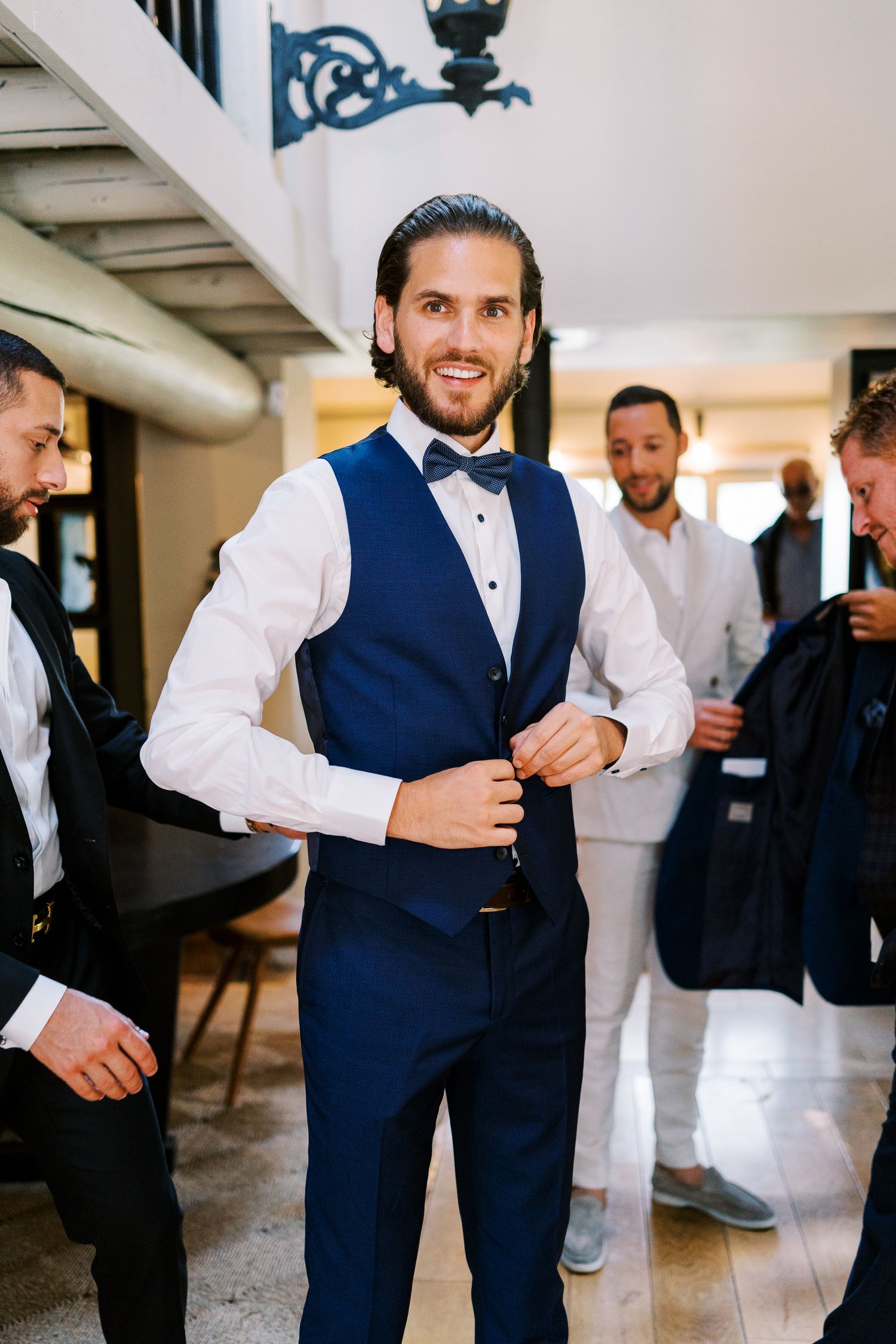Smiling groom in navy vest and bow tie buttoning his jacket while groomsmen prepare nearby.