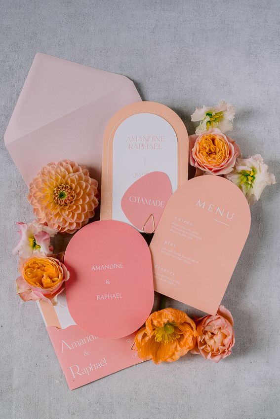 Pink and peach wedding stationery suite with arch-shaped menu cards, envelope, and orange floral accents on gray background.
