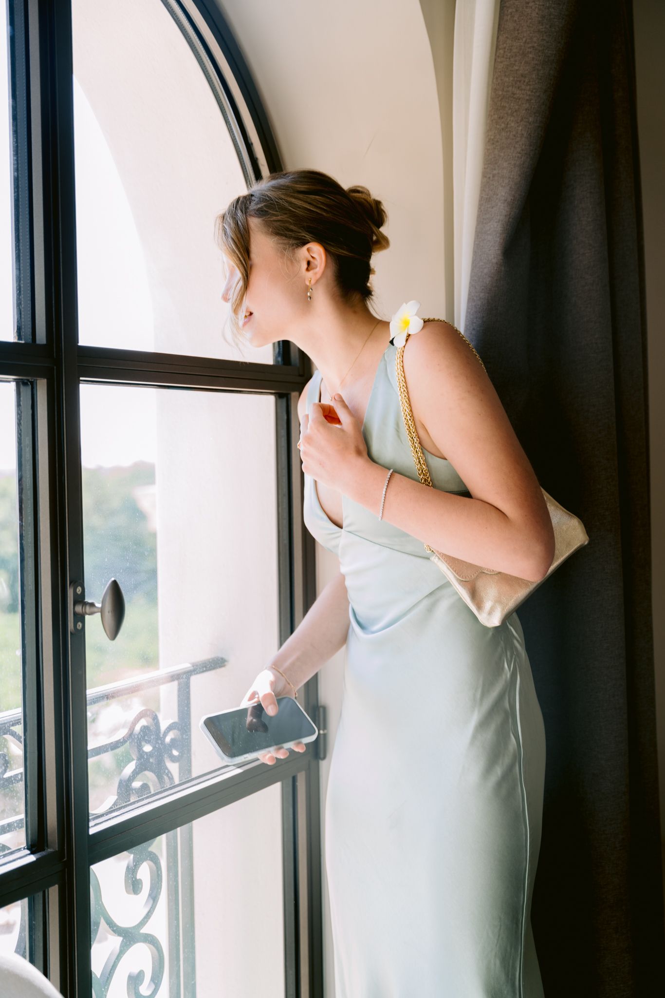 Person in white dress gazes through ornate windows while leaning on decorative balcony railing.