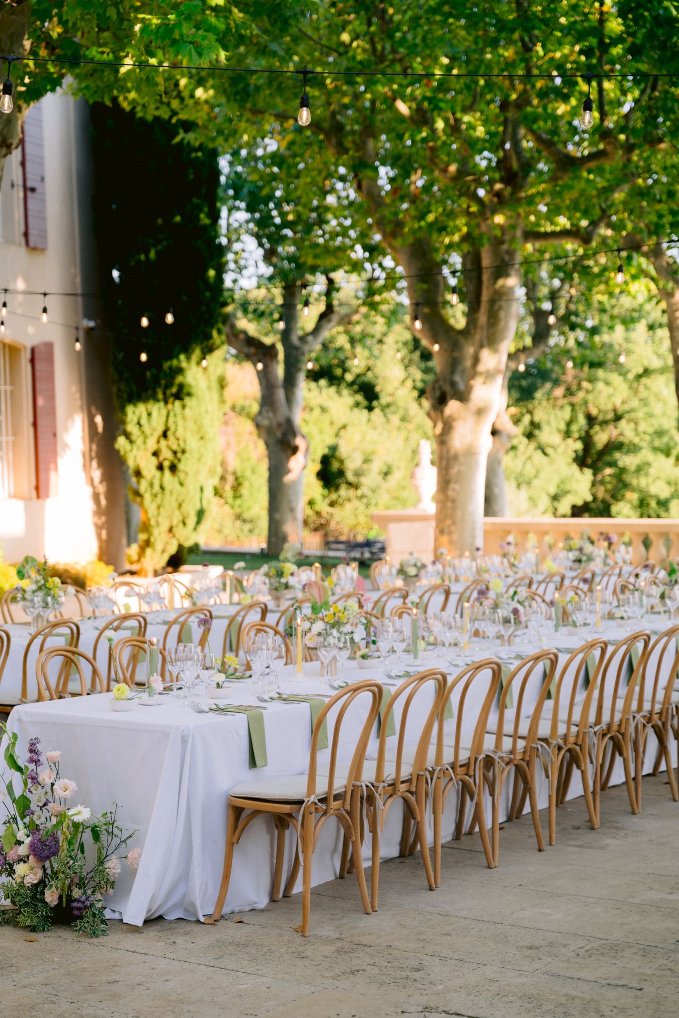 Long banquet tables with white linens and wooden cross-back chairs arranged under green trees for an outdoor event.