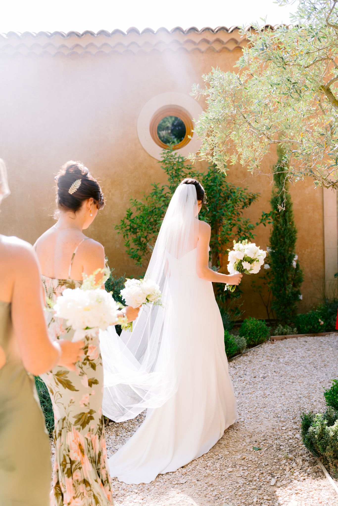 Bride in white gown and veil walks with bridesmaids in floral dresses, holding white bouquets in a sunlit garden.