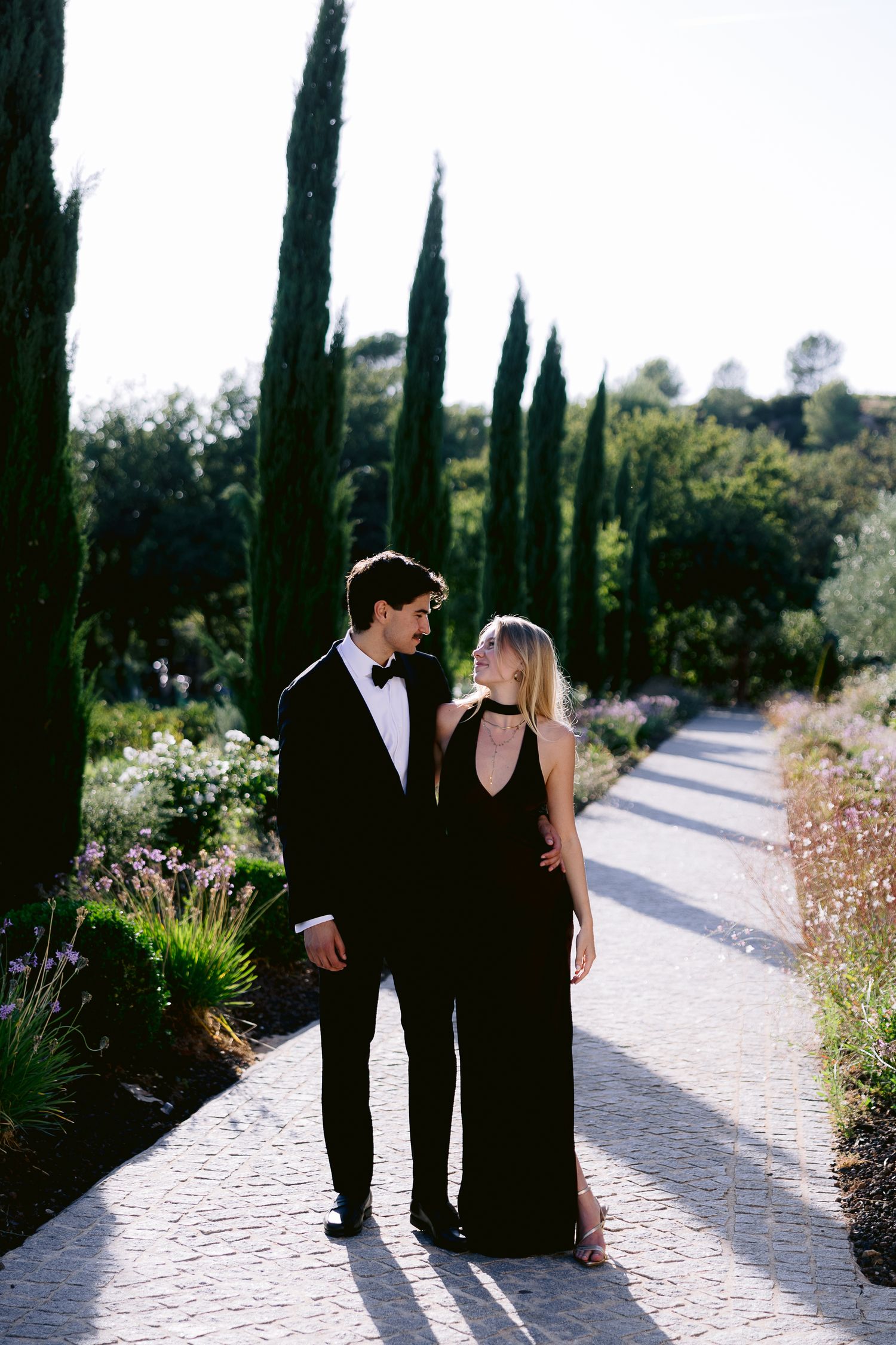 Elegant couple in black formal attire walking along a garden pathway lined with tall cypress trees.