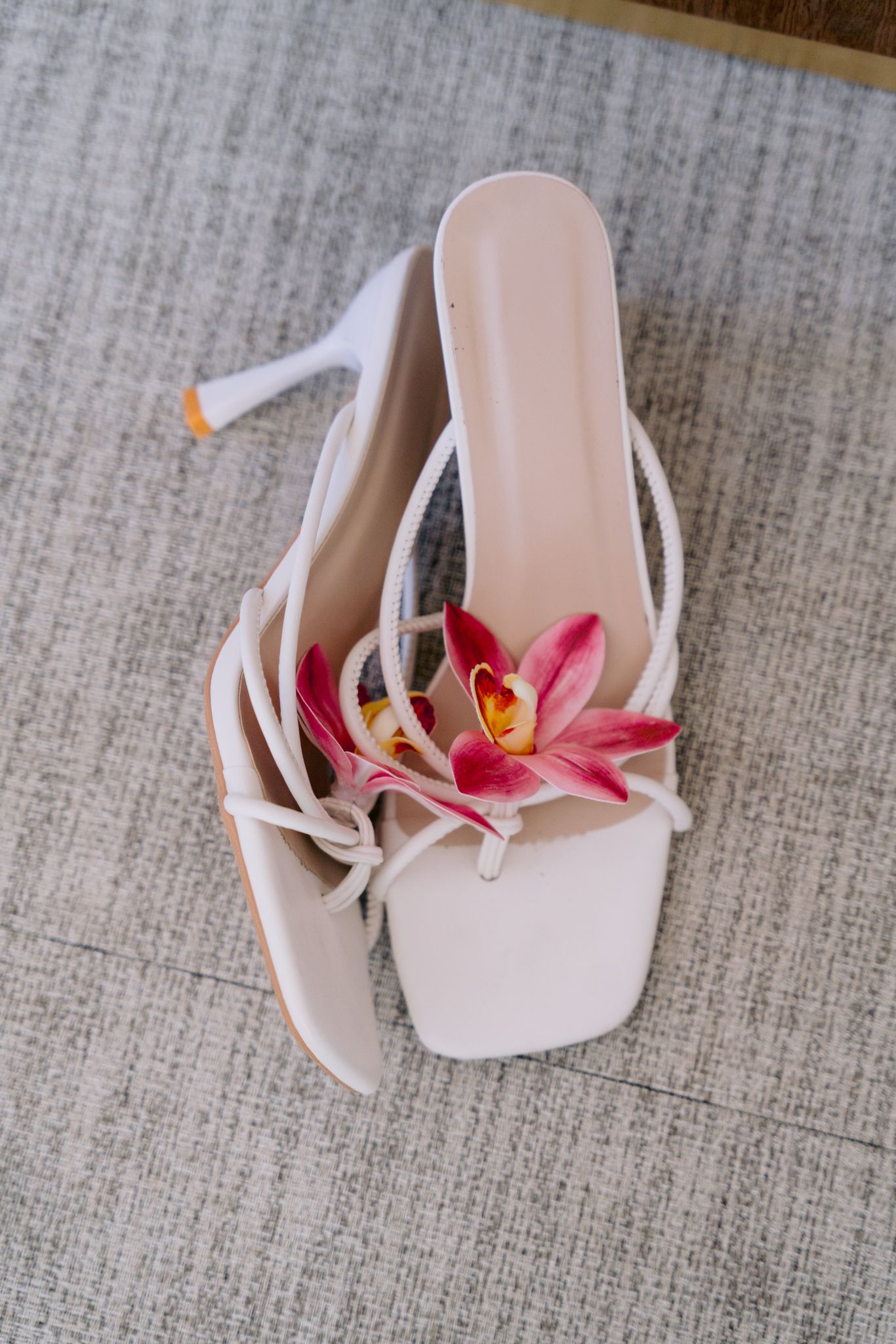 Pink orchid flower decoratively placed on elegant white leather sandals against a textured gray fabric background.