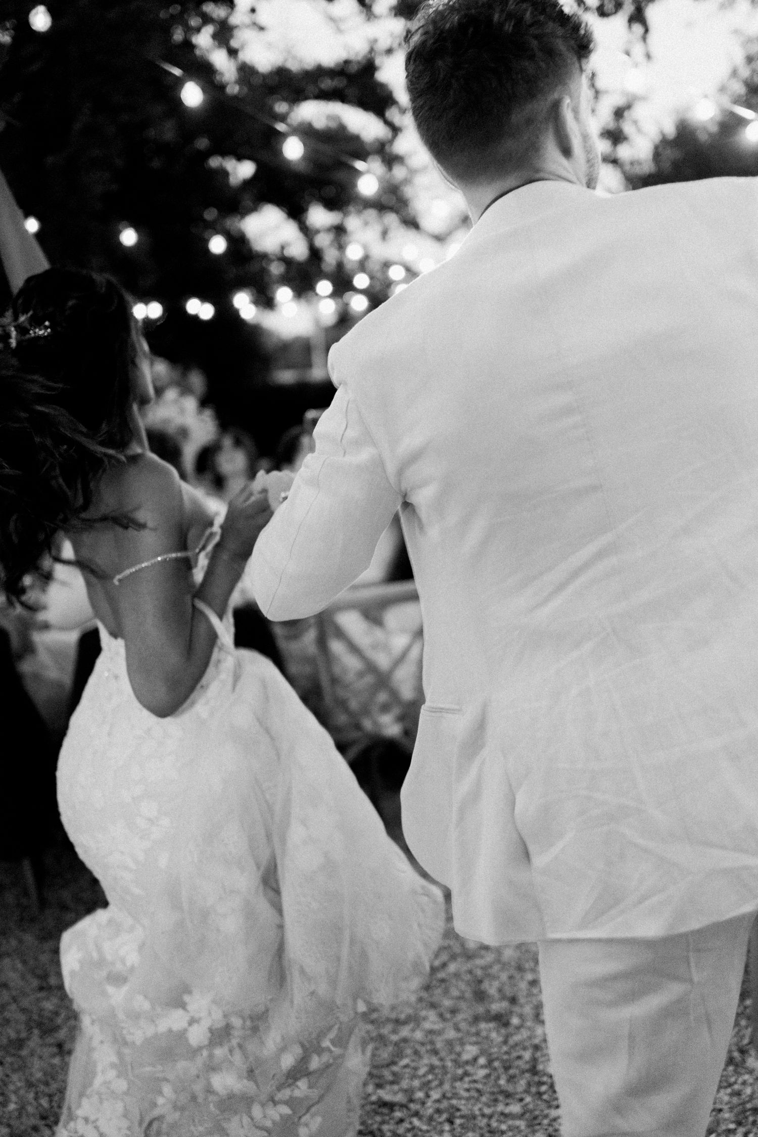 Black and white photo of bride and groom sharing their first dance outdoors under string lights at a wedding reception.