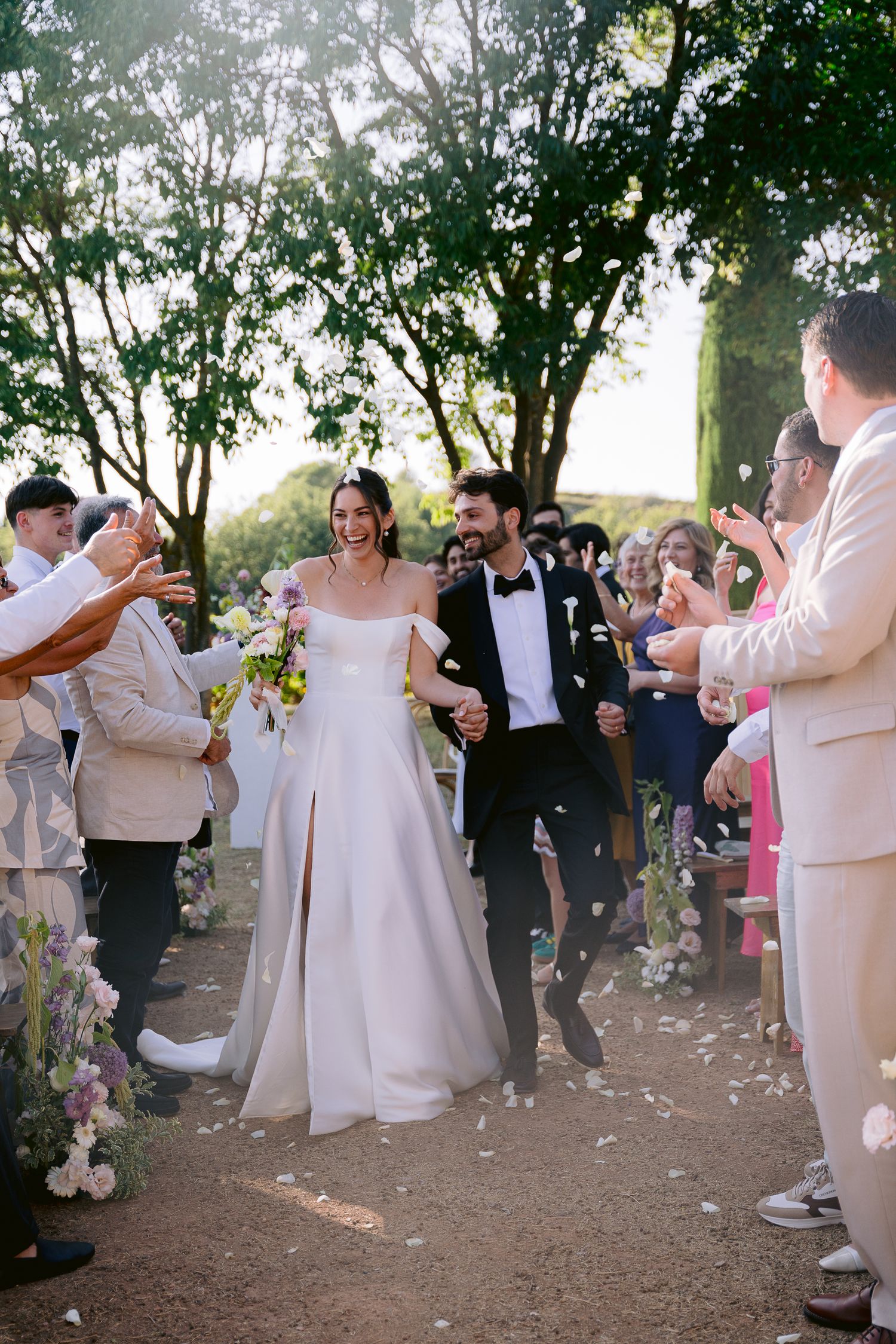 Bride and groom walk down outdoor aisle as guests toss white flower petals at a sunny garden wedding ceremony.
