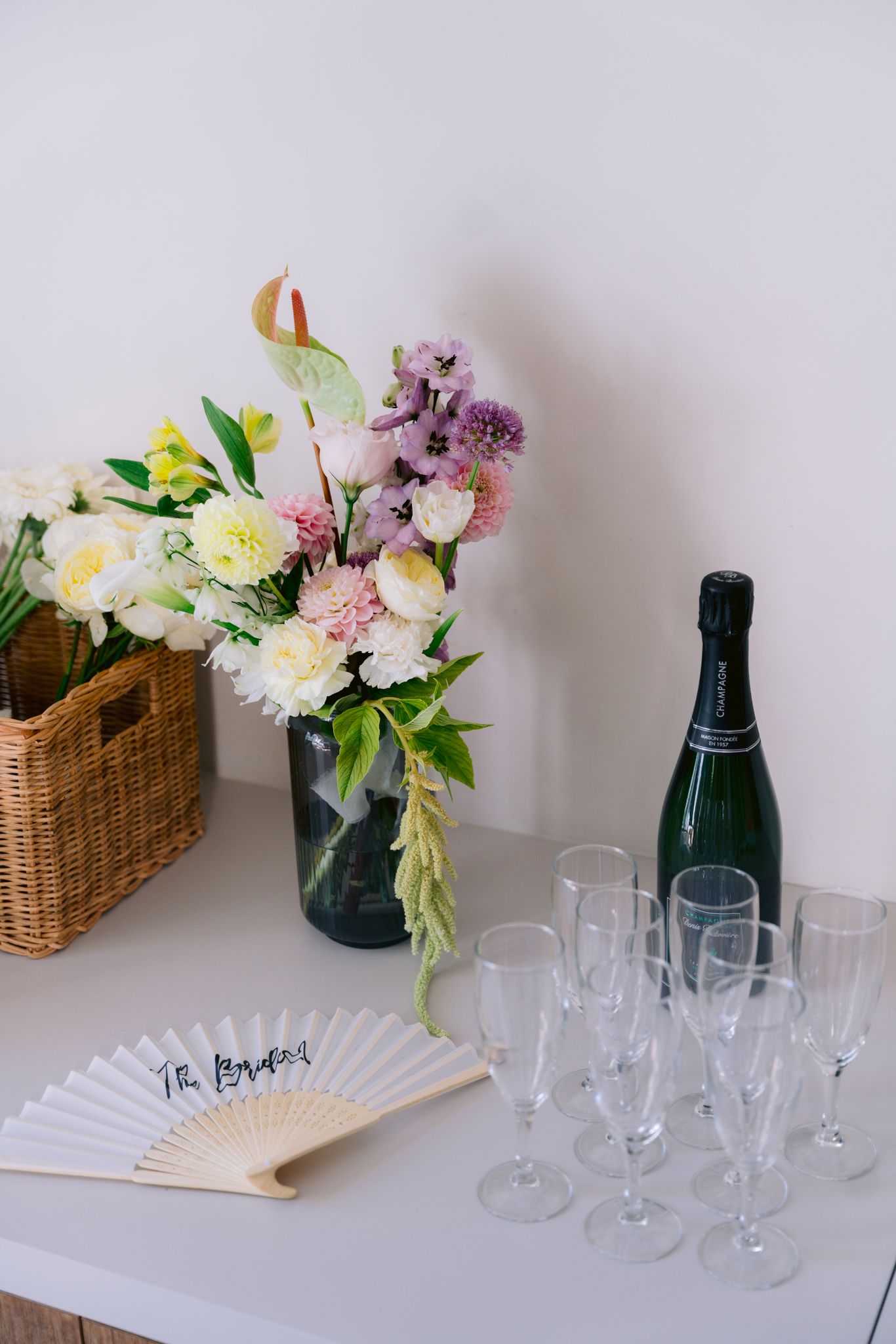 Elegant party setup with champagne flutes, paper fan, and fresh floral arrangement in blue vase on white table.