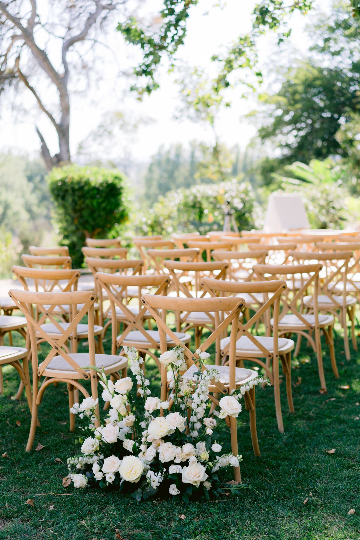 Outdoor wedding ceremony setup with wooden cross-back chairs on green lawn, adorned with white floral arrangements.