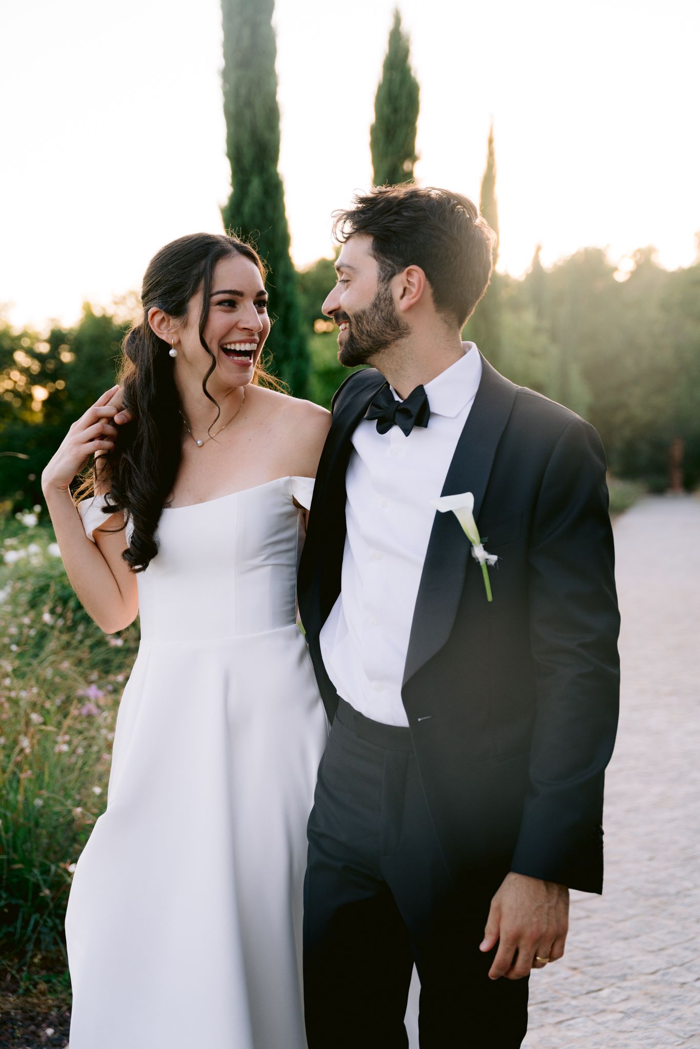 Bride in strapless white gown laughs joyfully with groom in black tuxedo at golden hour wedding.