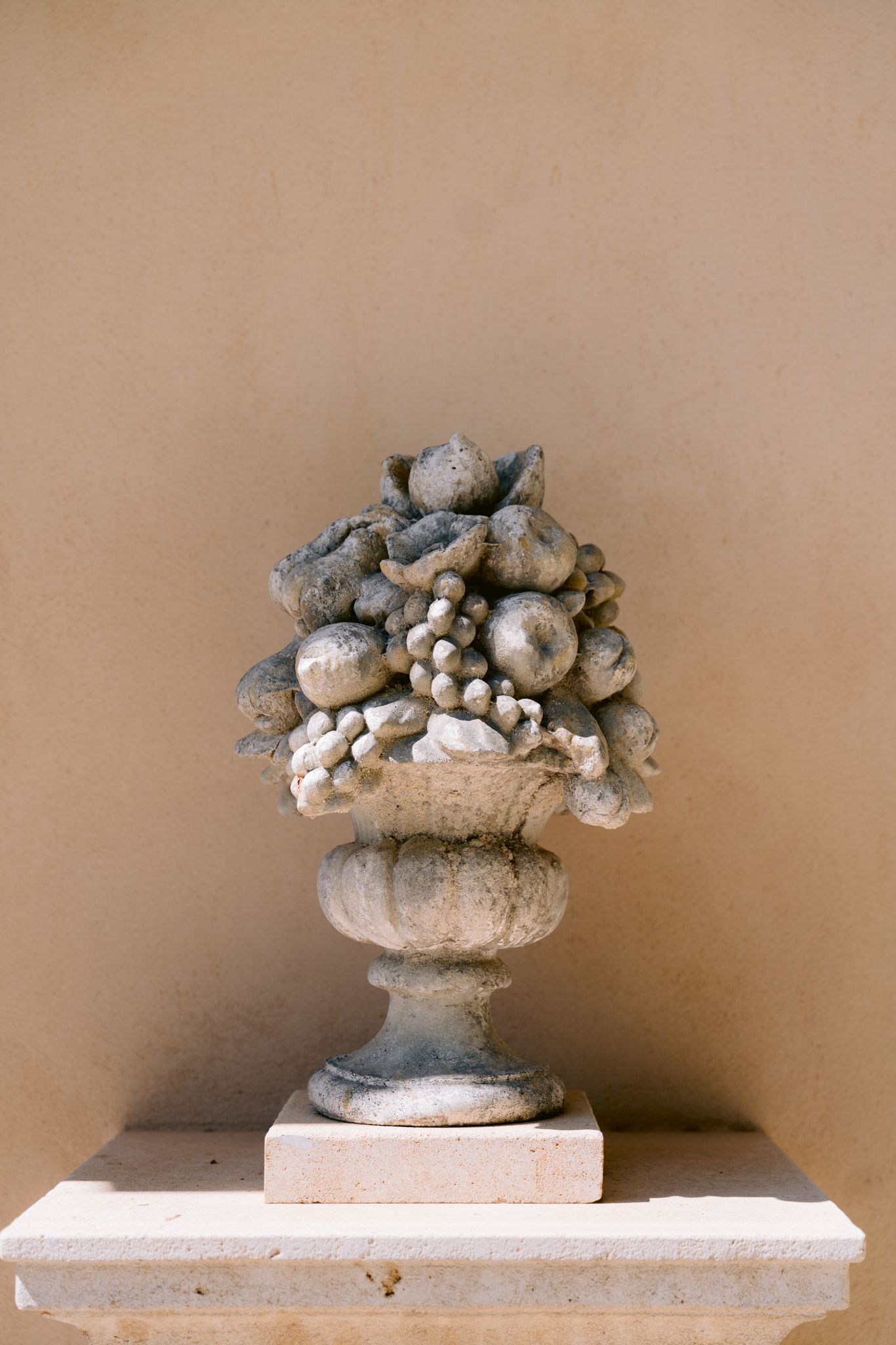 Decorative stone urns with fruit and floral designs displayed on white pedestals against a beige wall.