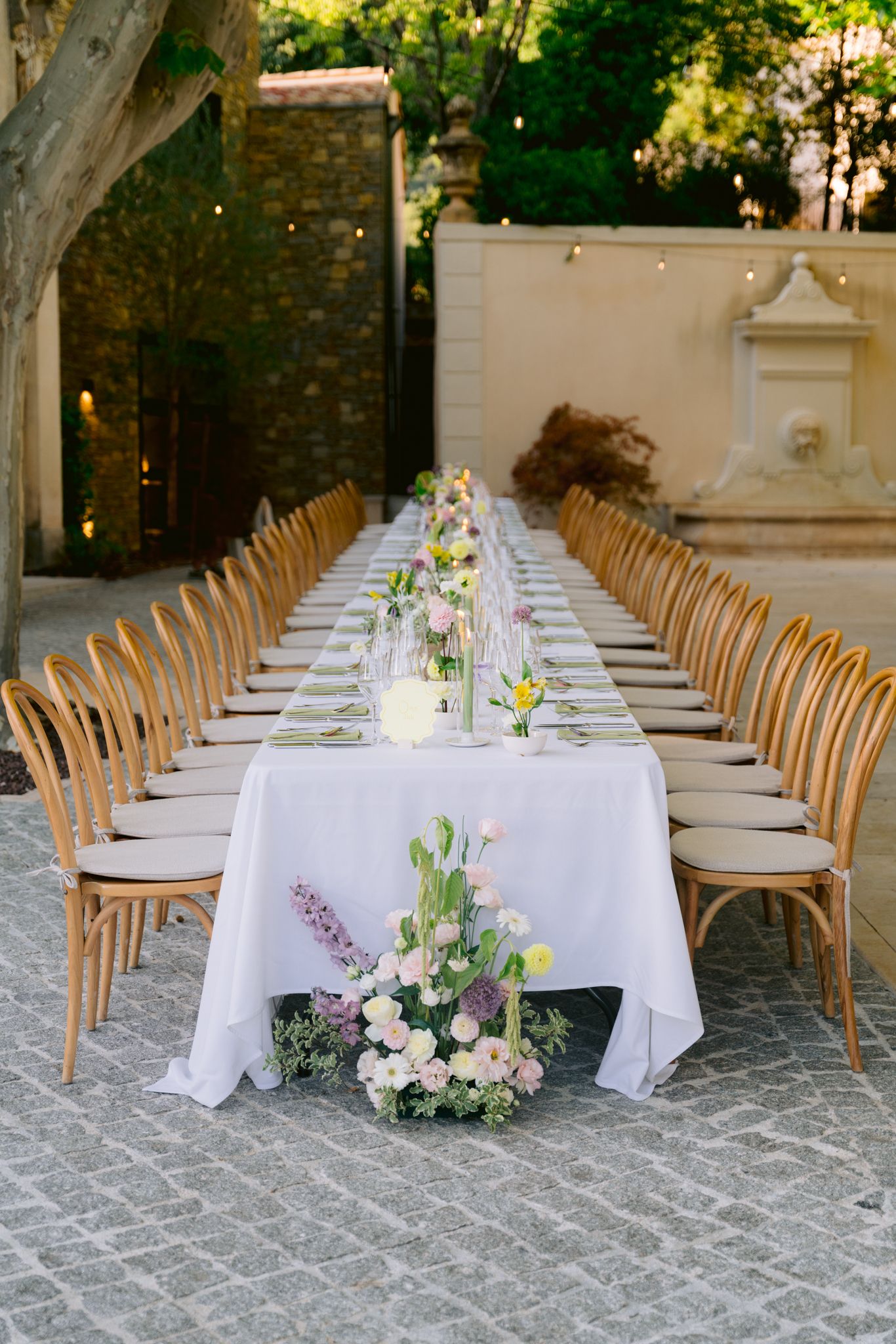 Long elegant outdoor dining tables with wooden chairs and white tablecloths decorated with fresh floral arrangements.