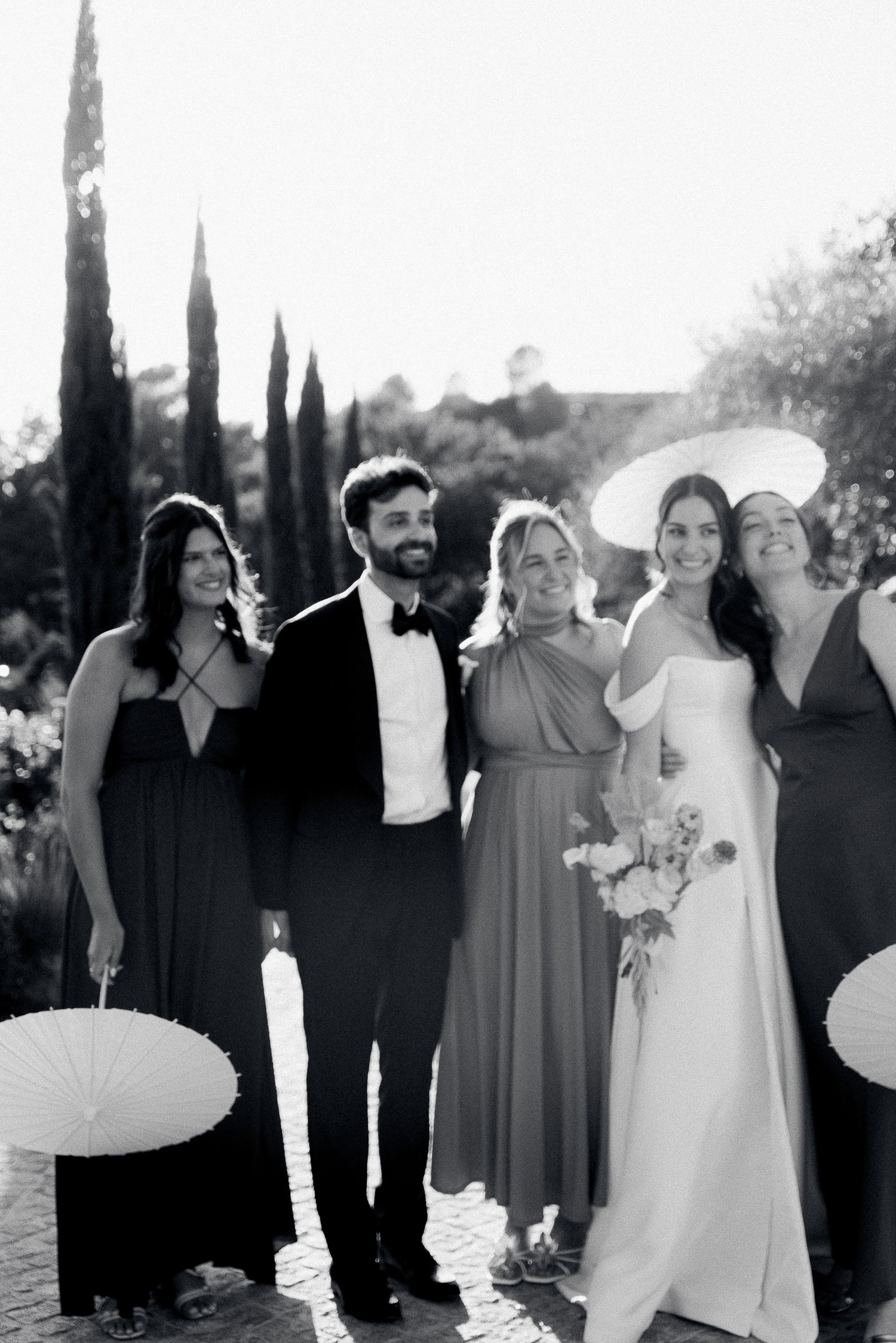 Black and white wedding photo of bride, groom, and bridesmaids posing outdoors with parasols and cypress trees.