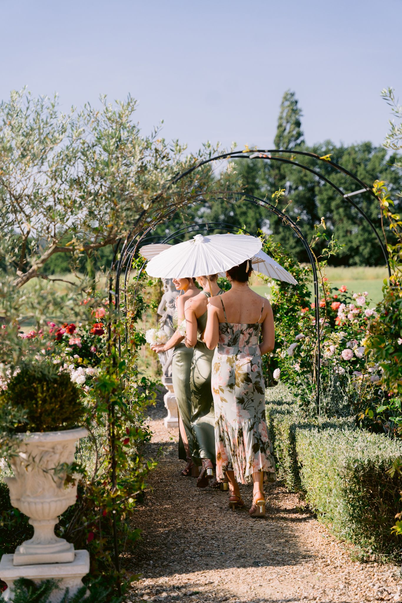 Bridesmaids in floral gowns walk through a rose garden arch carrying white parasols.