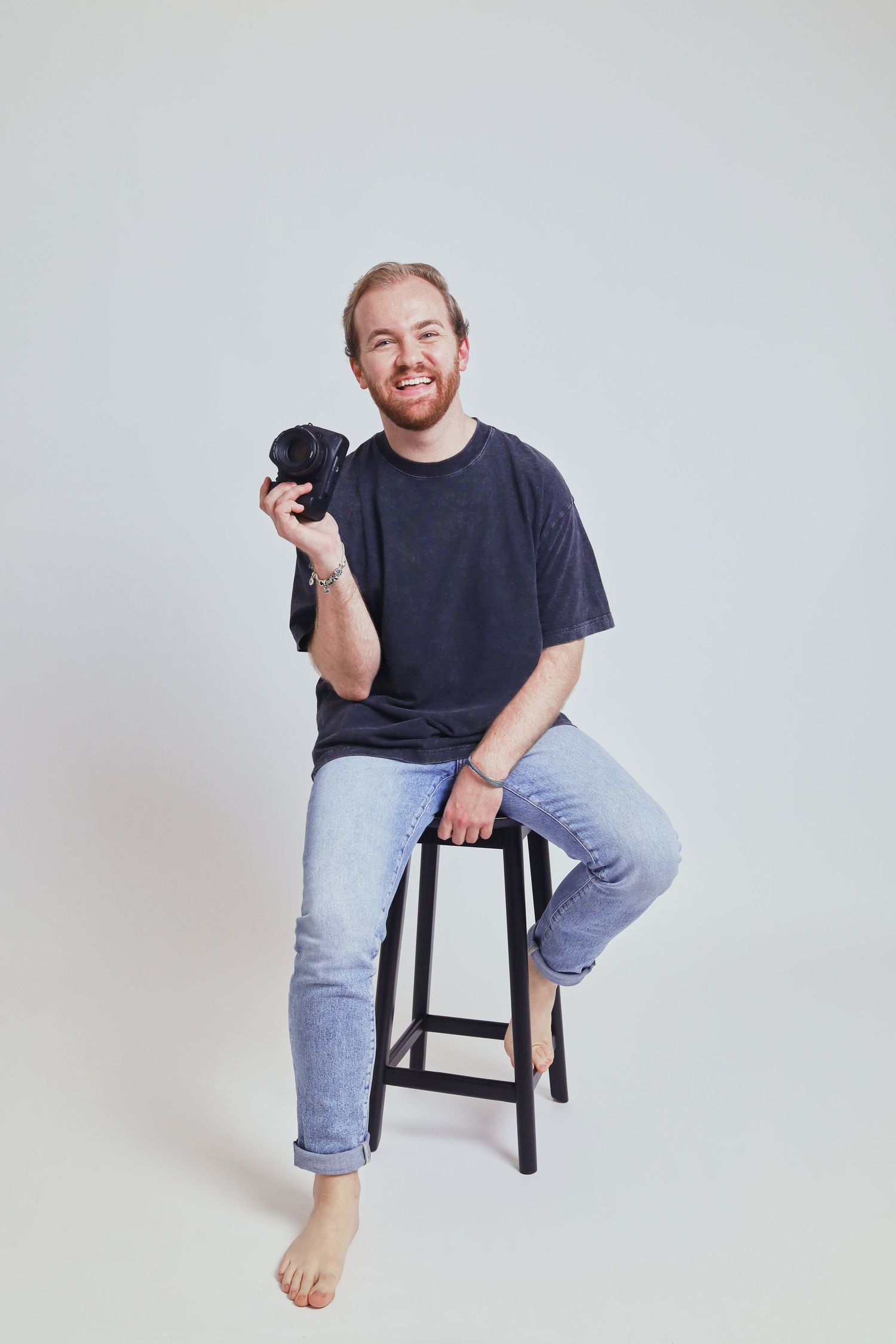 Smiling man sitting on a stool holding a camera, wearing a dark t-shirt and jeans, barefoot against a white background.