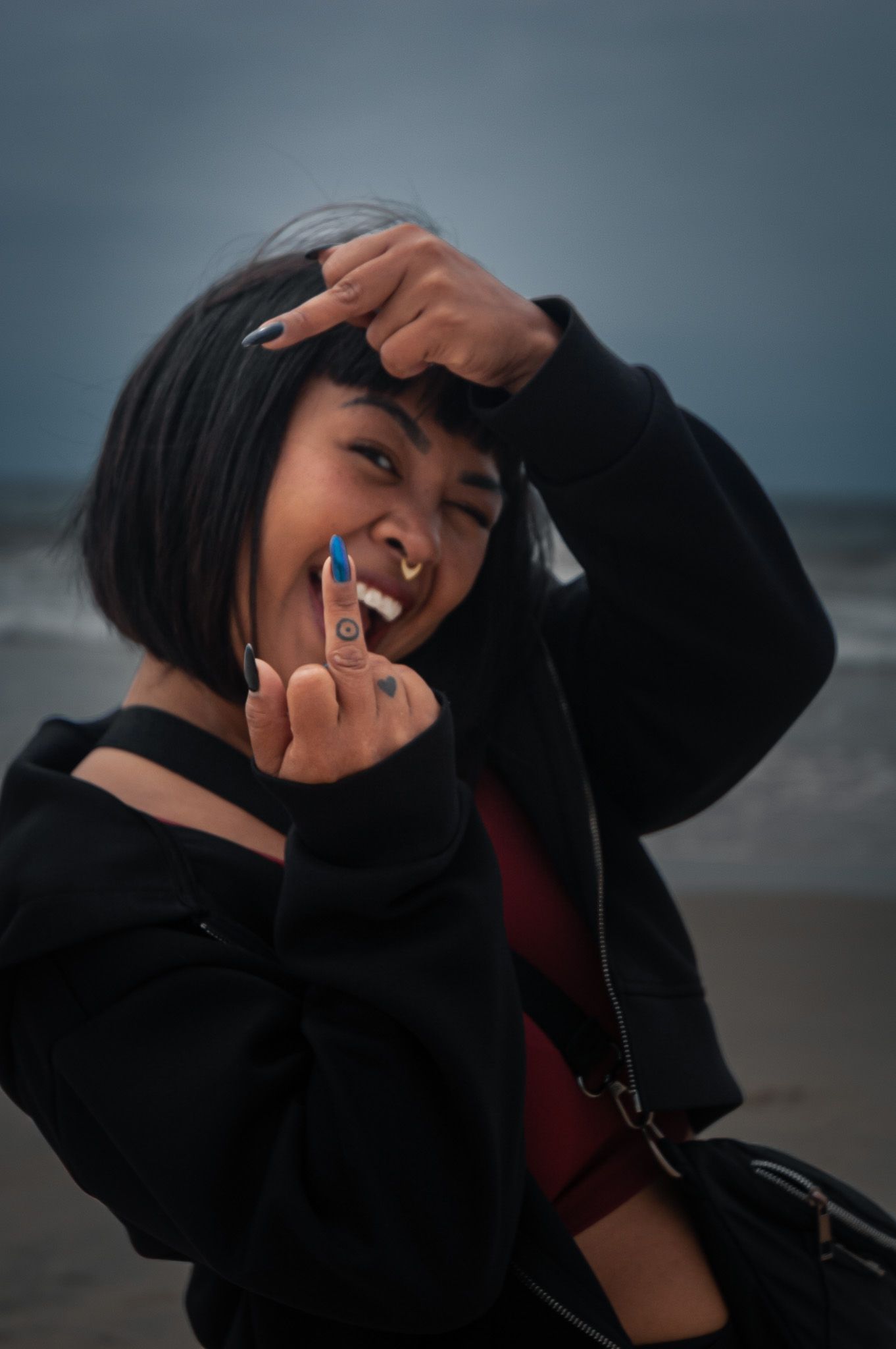 A person in a black sweater smiling playfully against an overcast sky at the beach.