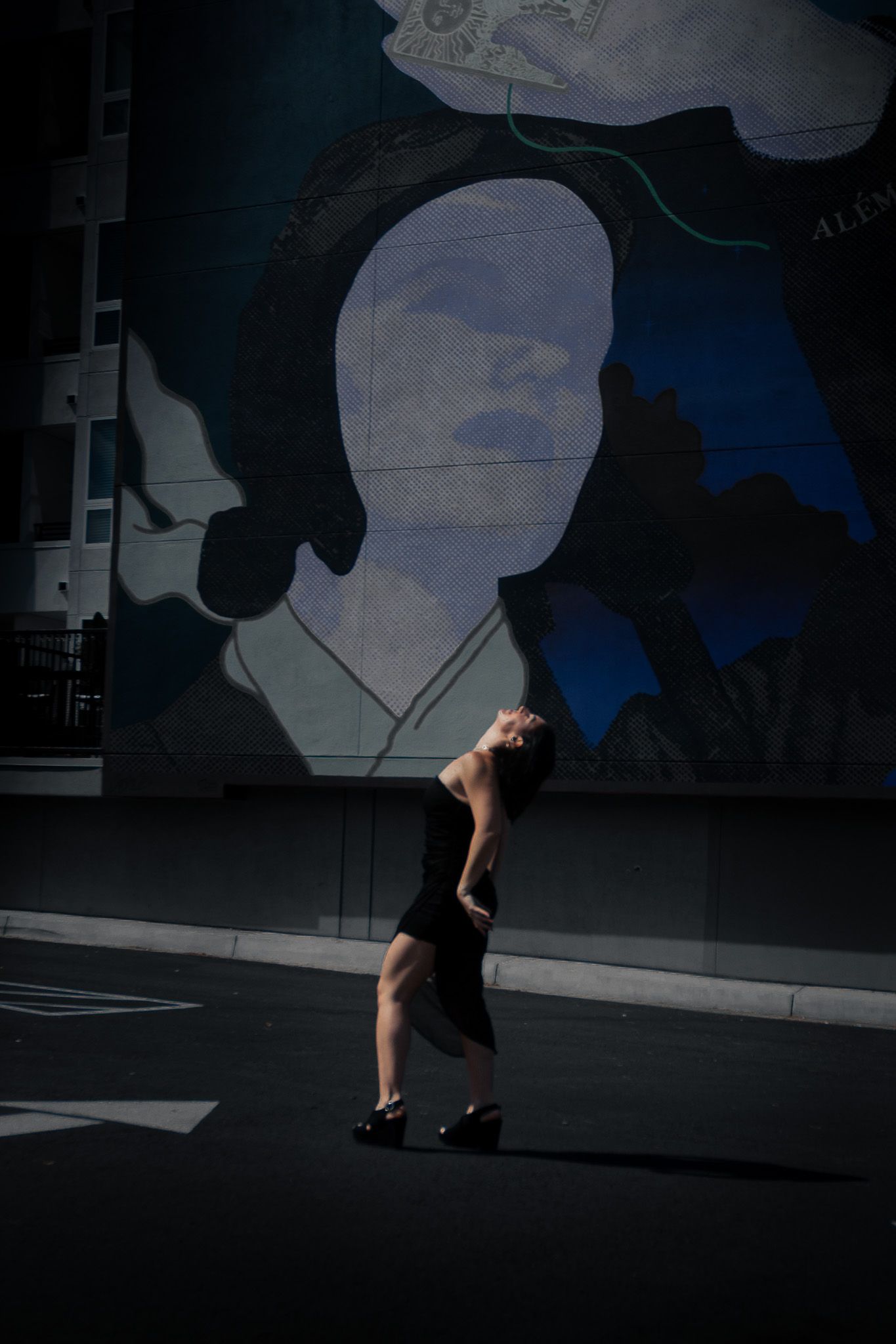 A dancer in a black dress strikes a graceful pose against a large painted mural backdrop on an urban street at dusk.