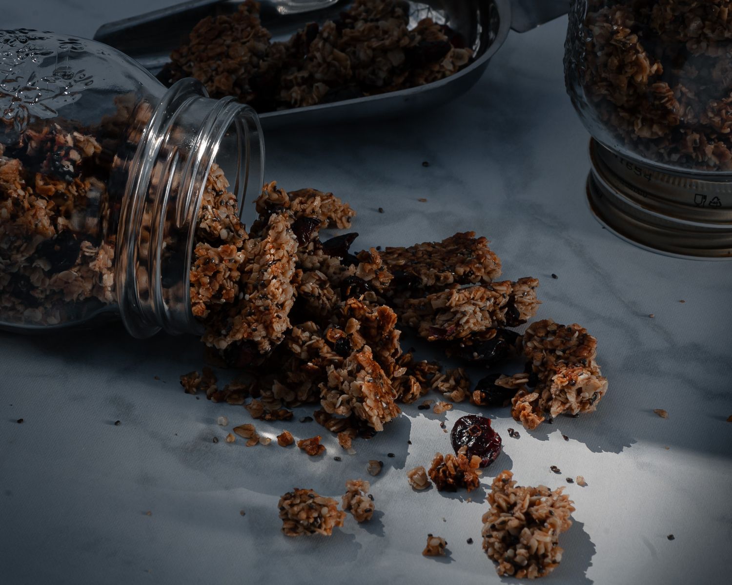 Dark chocolate granola clusters spilling out of a glass mason jar onto a gray surface in moody lighting.