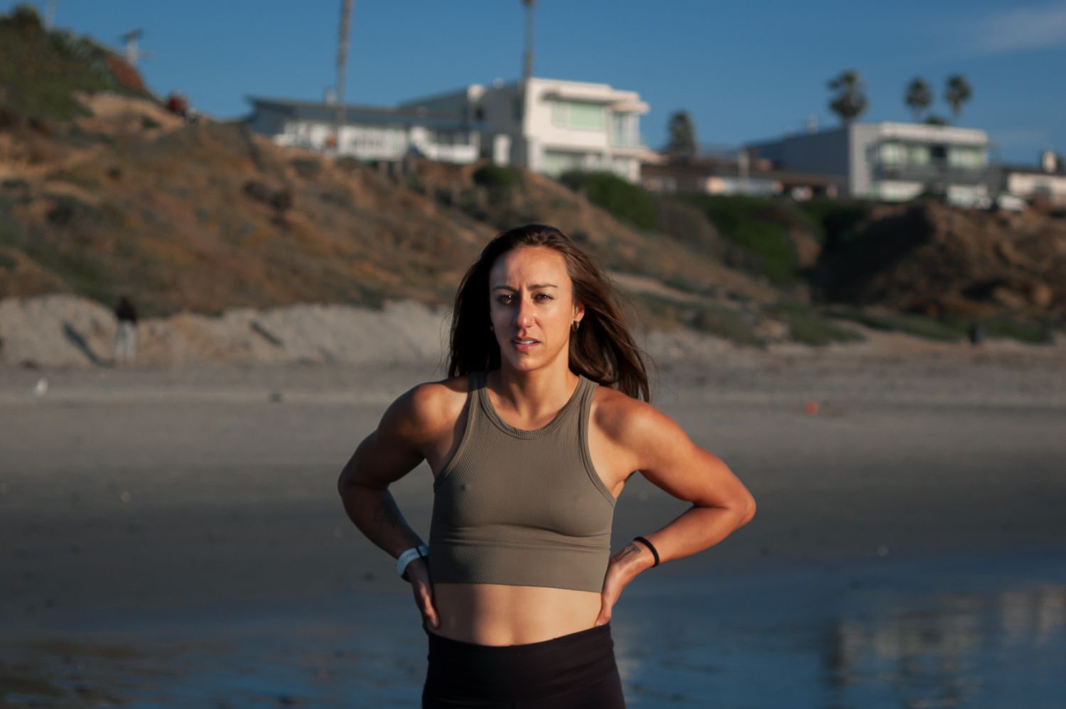 Athletic person in workout attire stands with hands on hips during coastal beach workout at sunset.