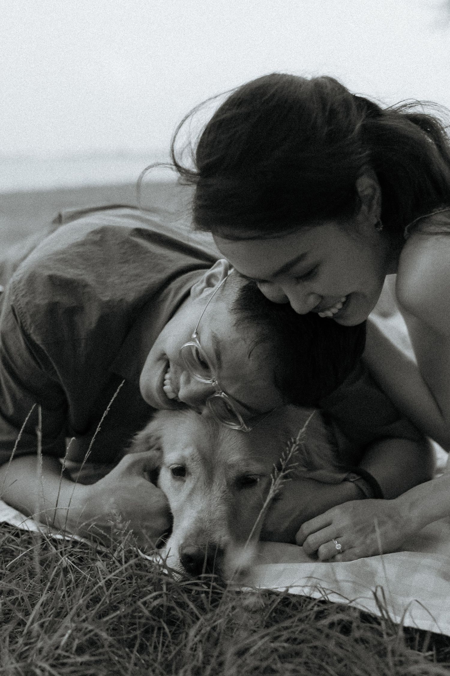 Black and white photo of smiling couple lying down cuddling with their golden retriever.