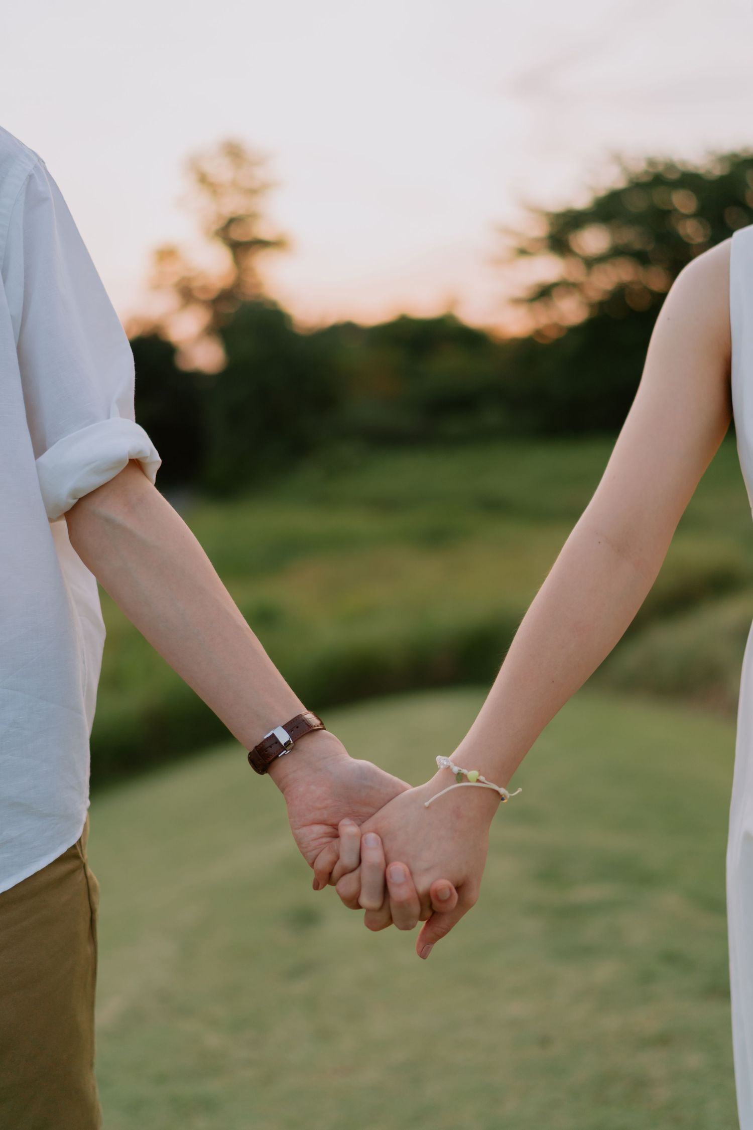 Close-up of a couple holding hands at sunset, showcasing their matching bracelets outdoors.