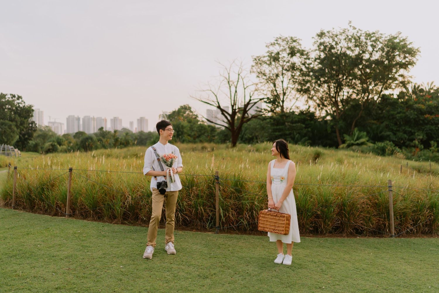 Couple standing apart in a lush meadow at sunset, man holding camera, woman carrying wicker basket.