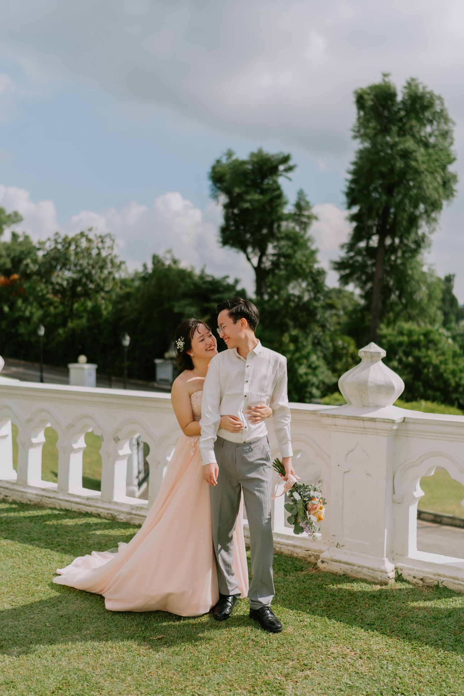 Bride in pink gown leaning on groom beside white balustrade with lush green trees behind them.