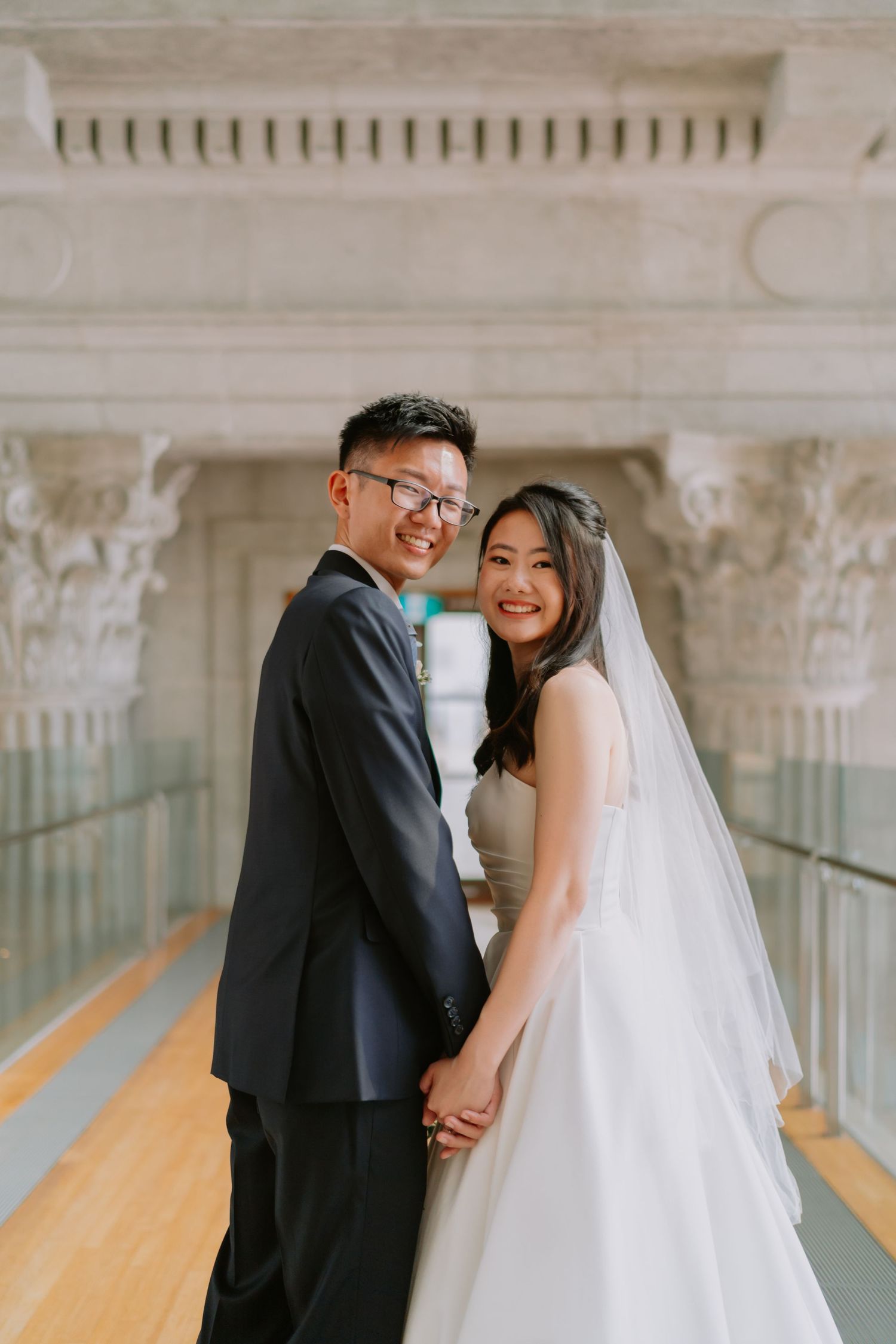 Smiling Asian bride and groom holding hands in front of elegant marble columns.