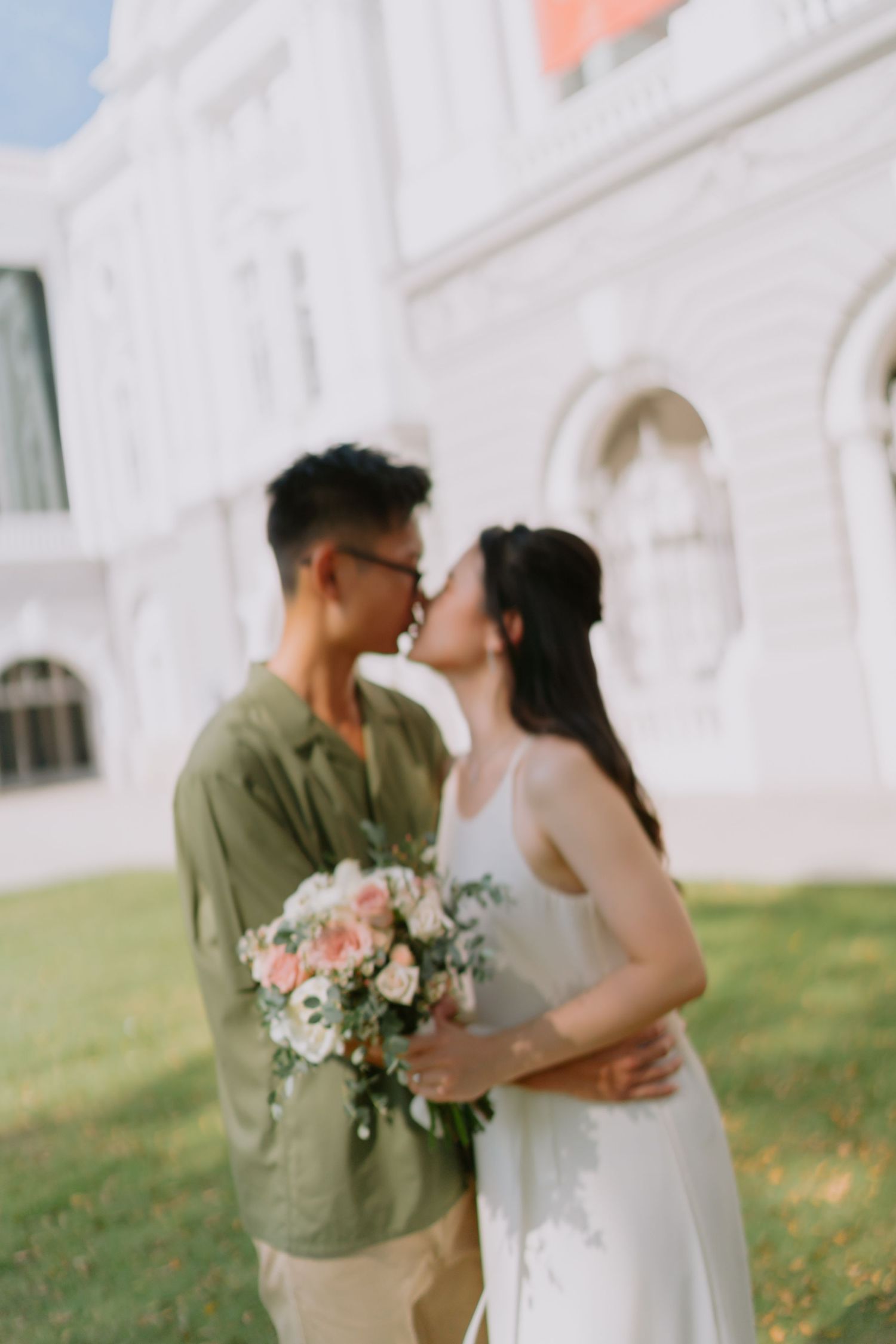 Couple sharing a kiss on lawn in front of white colonial building with floral bouquet.