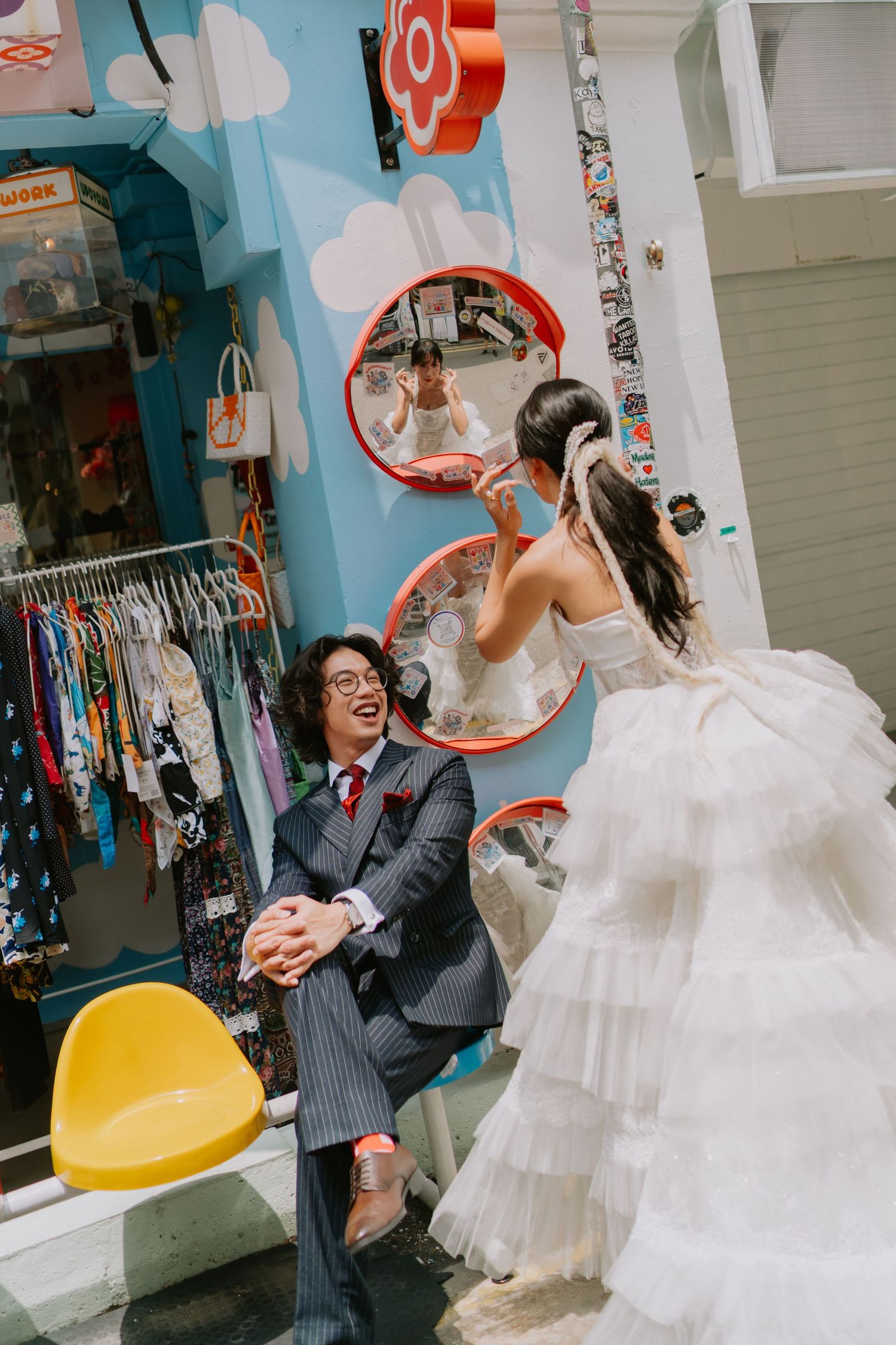 Laughing groom kneeling before bride outside colorful boutique with circular mirror reflections.