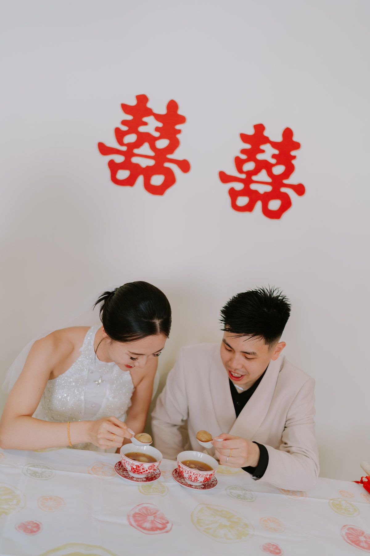 Couple in wedding attire sharing tea ceremony with red double happiness symbols on white wall behind them.
