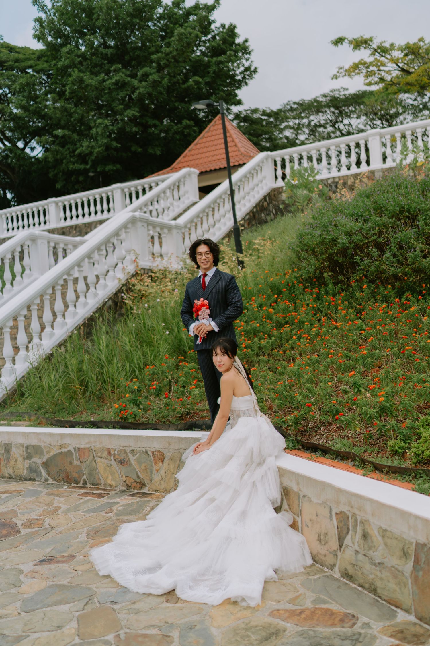 Elegant bride in layered white gown and groom with bouquet posing on stone steps surrounded by lush greenery.