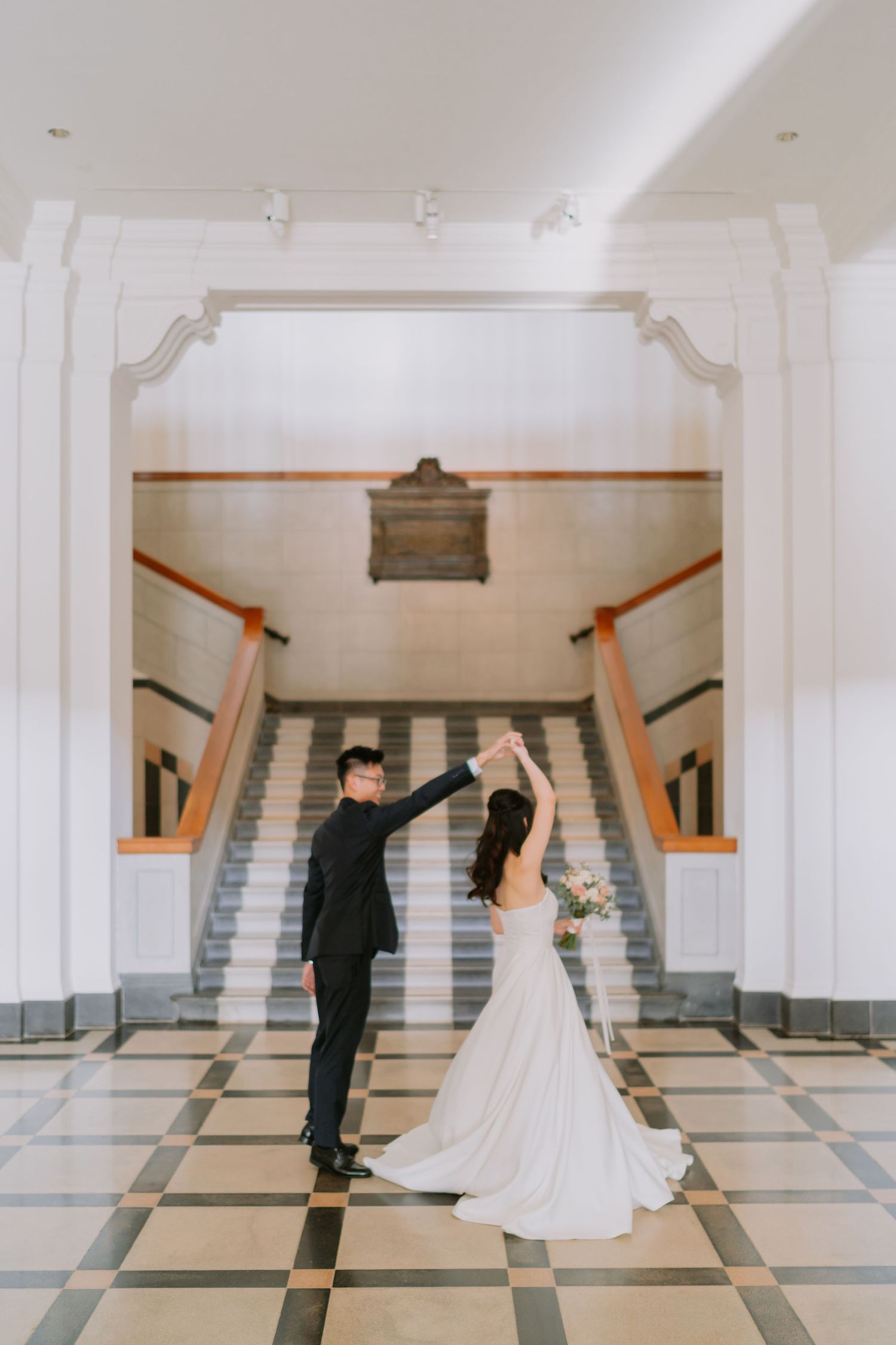 Bride and groom dancing in elegant white marble hall with checkered floors and grand staircase.