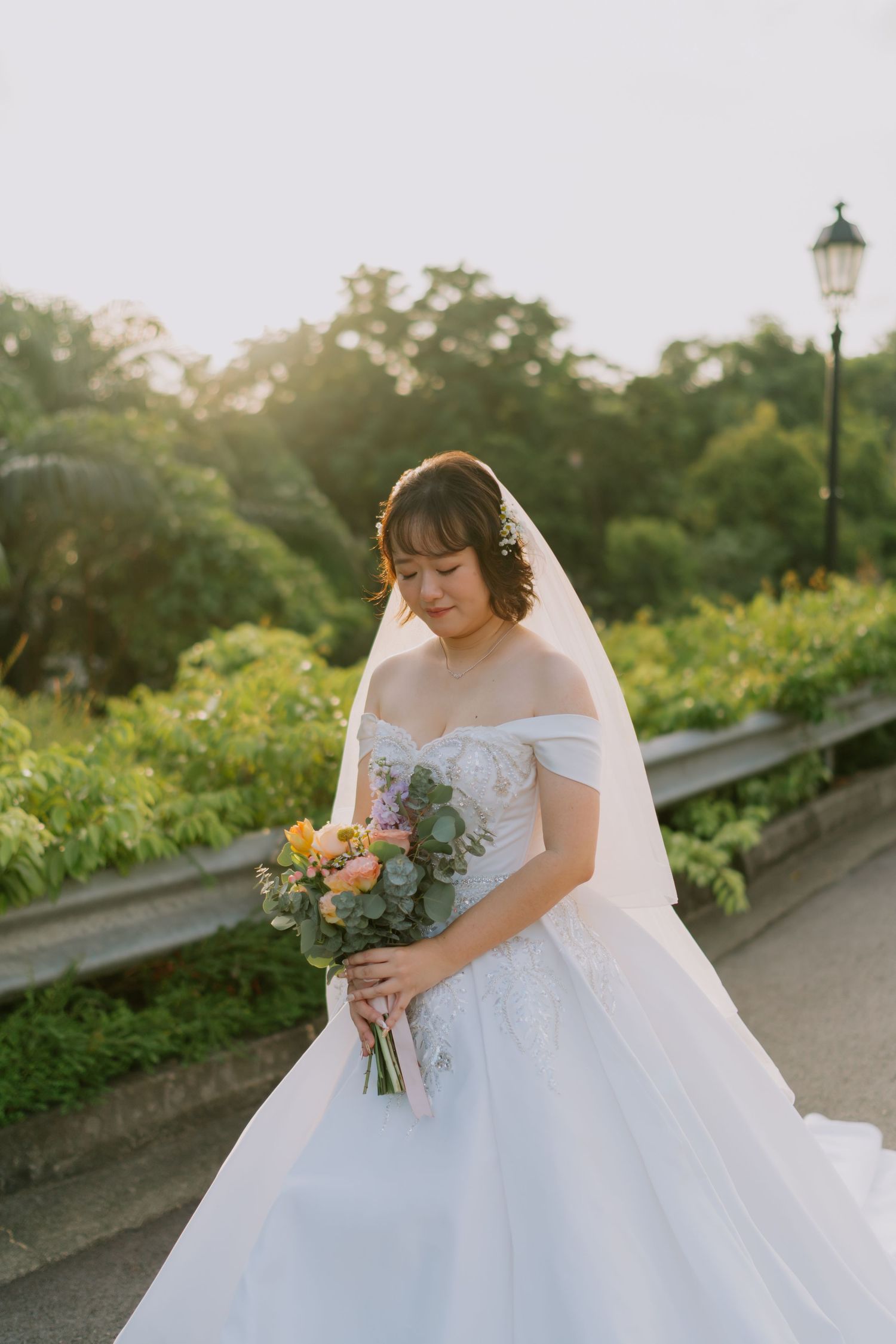 Asian bride in white off-shoulder gown gazes down at colorful bouquet in warm sunset light.