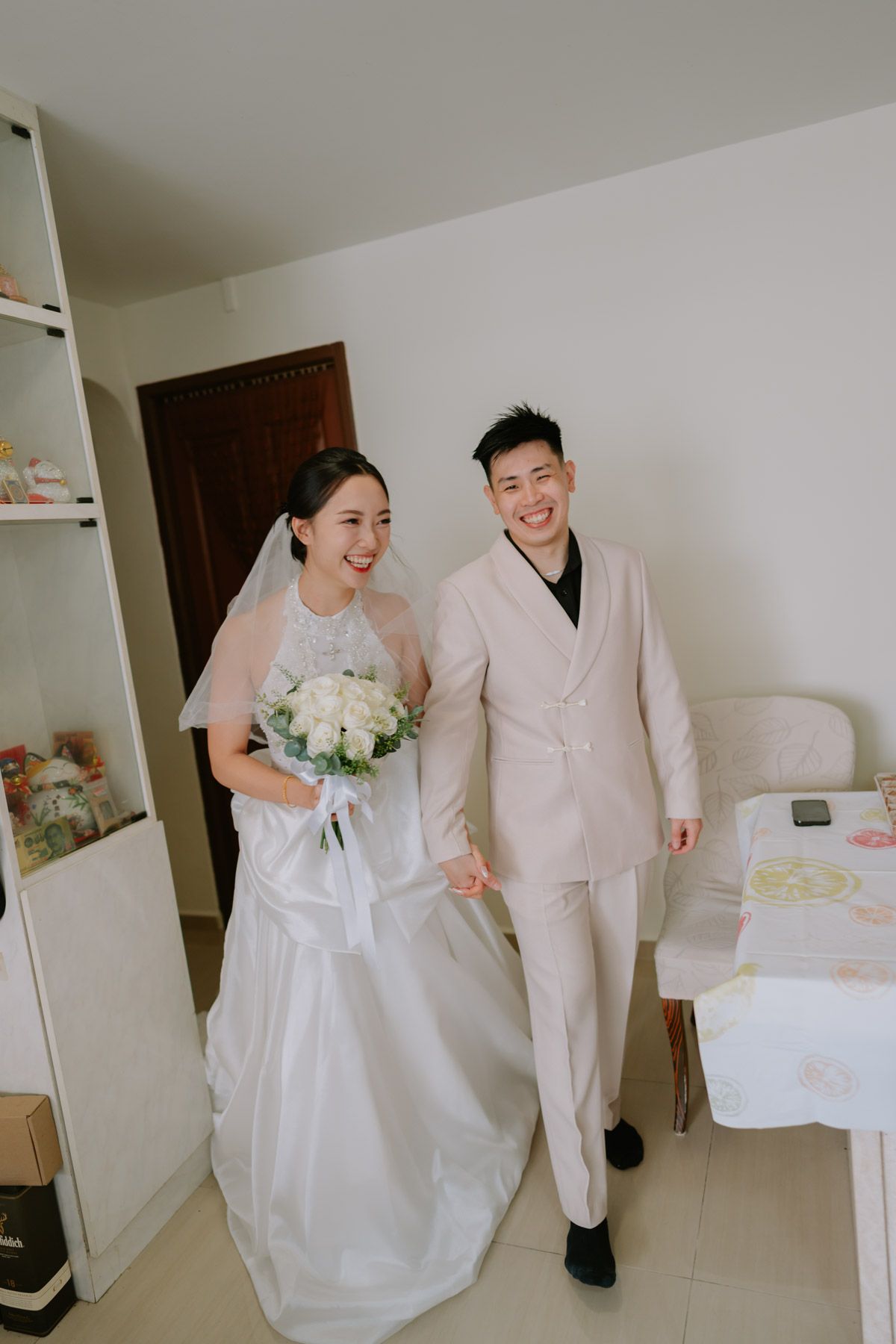Happy Asian couple in wedding attire, bride in white gown with bouquet, groom in cream suit, walking indoors.