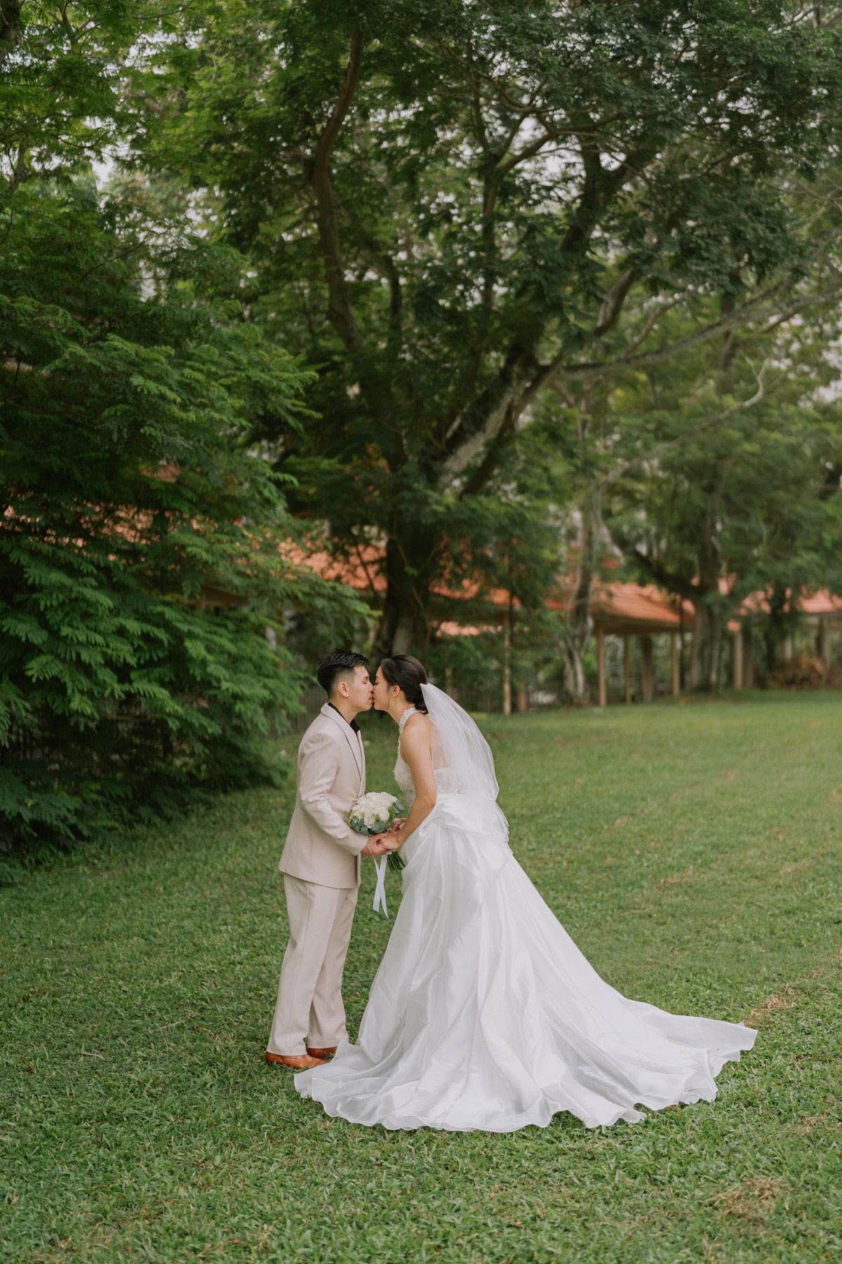 A couple shares a romantic kiss in a lush green garden, bride in a flowing white gown holding a bouquet, groom in a beige suit.