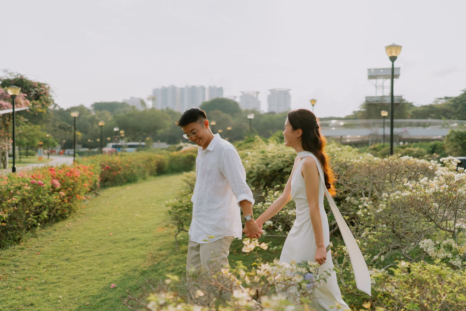 Couple holding hands walking through a garden path with colorful flowers and city skyline.
