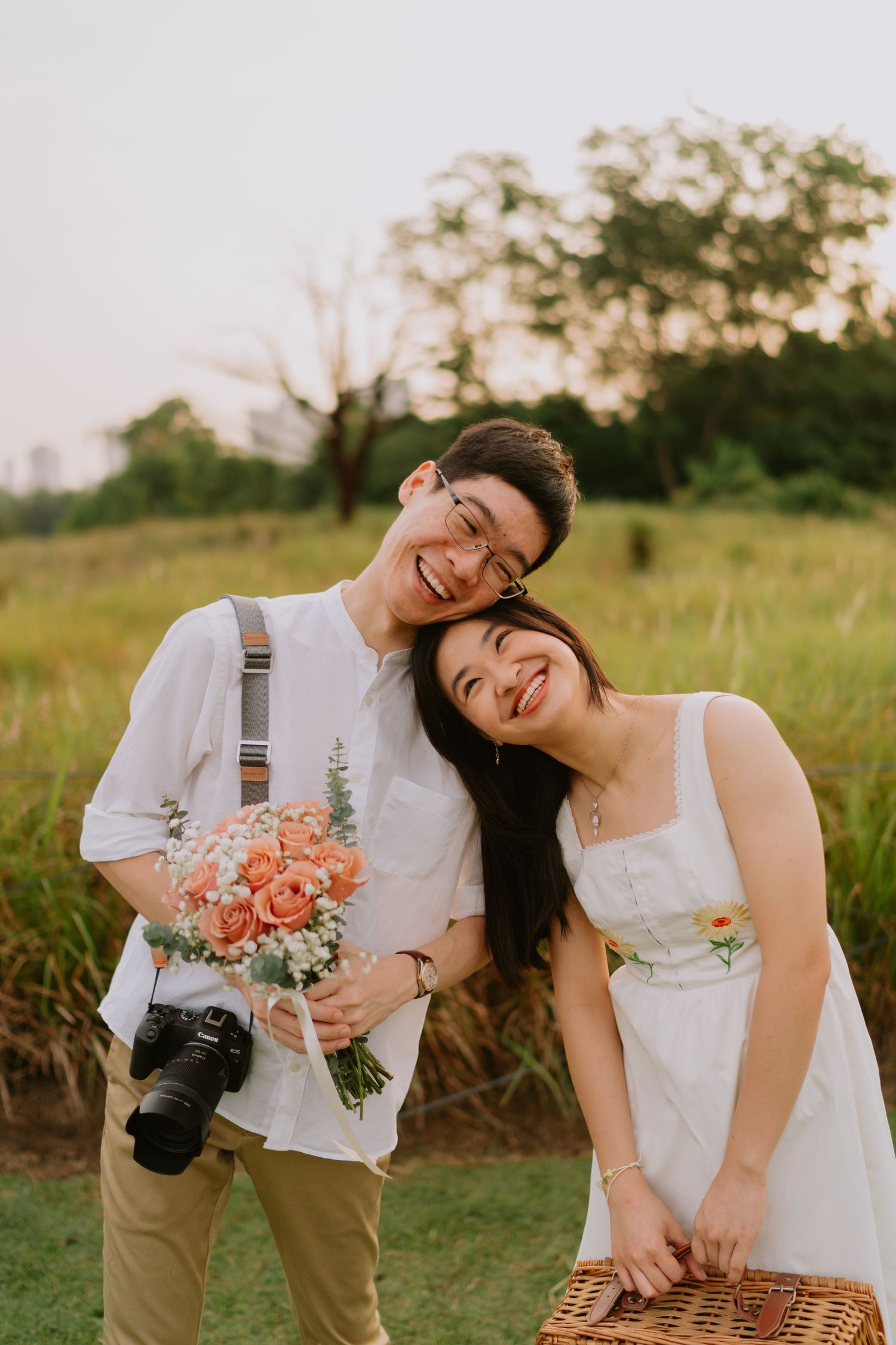 Happy Asian couple smiling together outdoors, man with camera and flowers, woman in white dress.