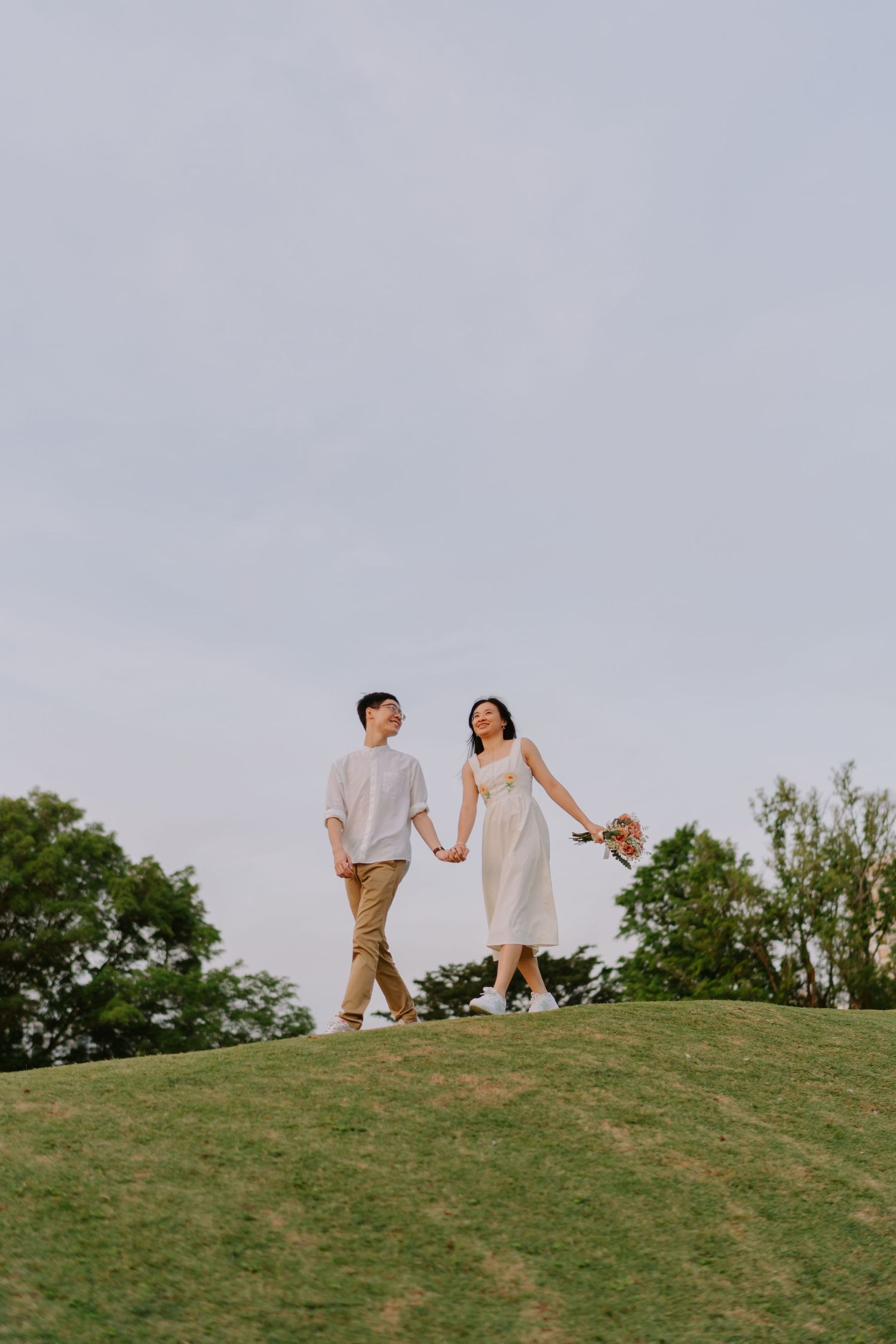 A couple holds hands while walking on a grassy hill, the woman in a white dress holding a bouquet.