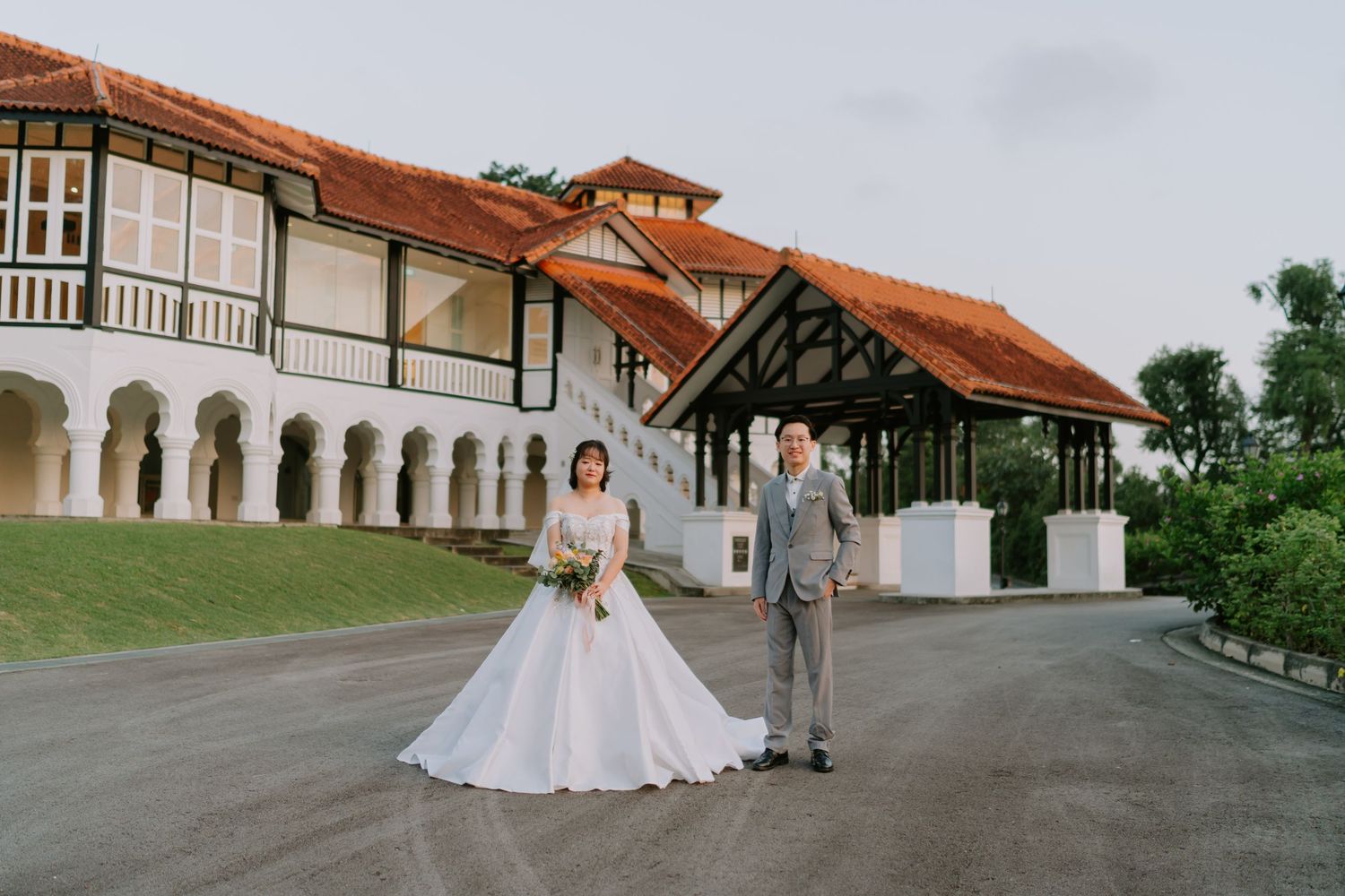 A bride in a white gown and groom in gray suit pose before an elegant colonial building.
